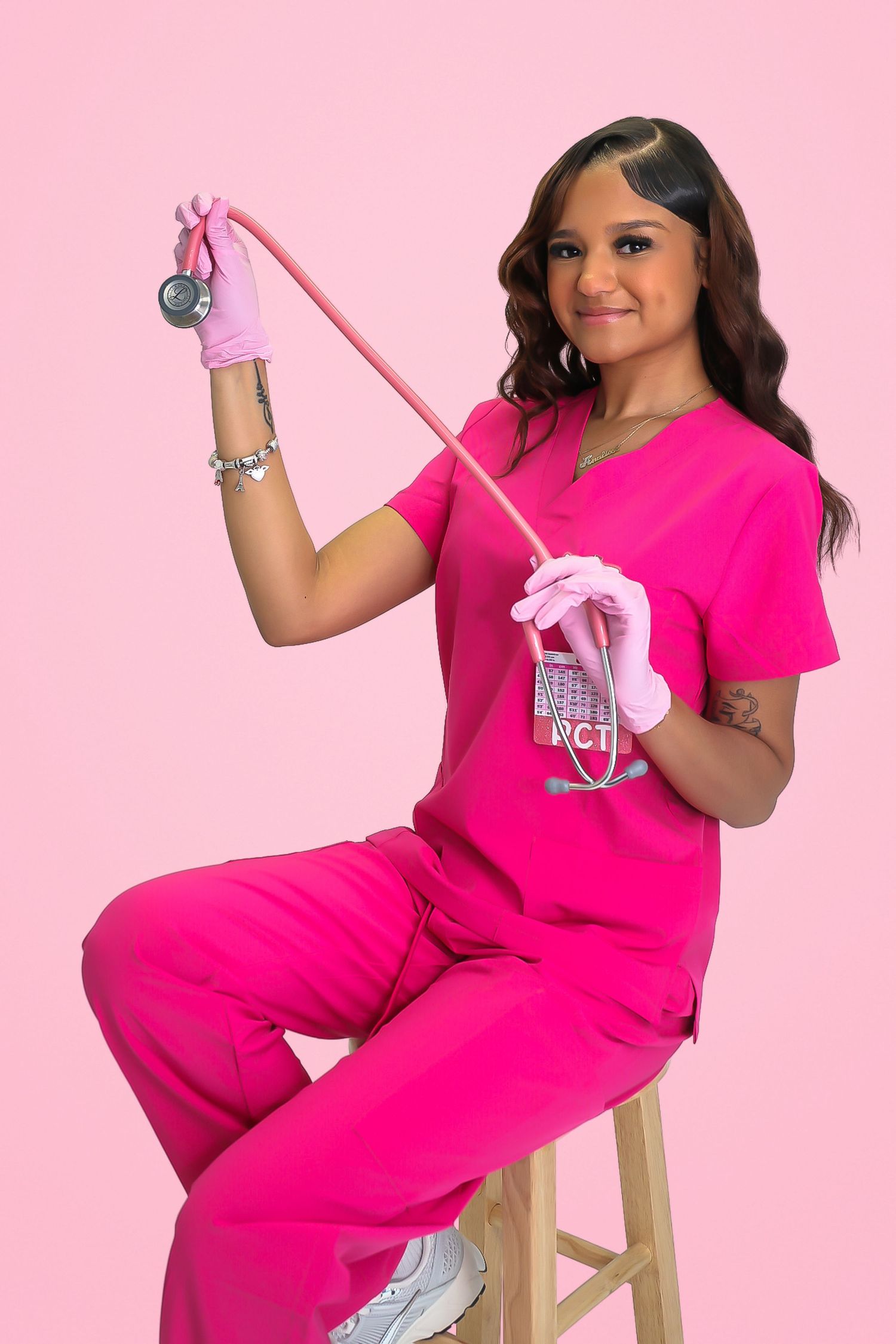 A smiling nurse in pink scrubs and gloves holds a stethoscope while seated on a stool against a pink background.