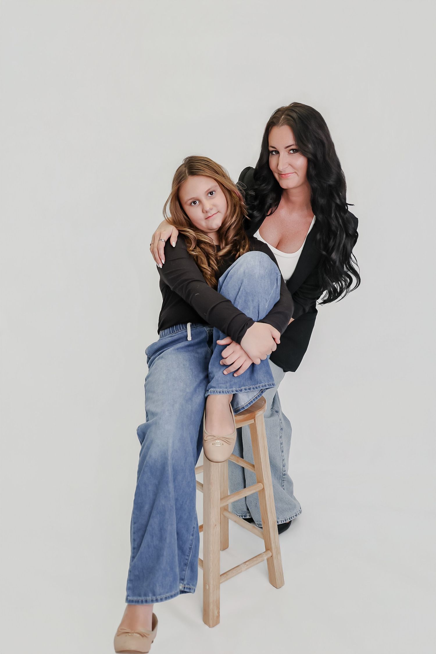 A woman and young girl in matching black jackets and jeans pose together on a wooden stool against a white background.