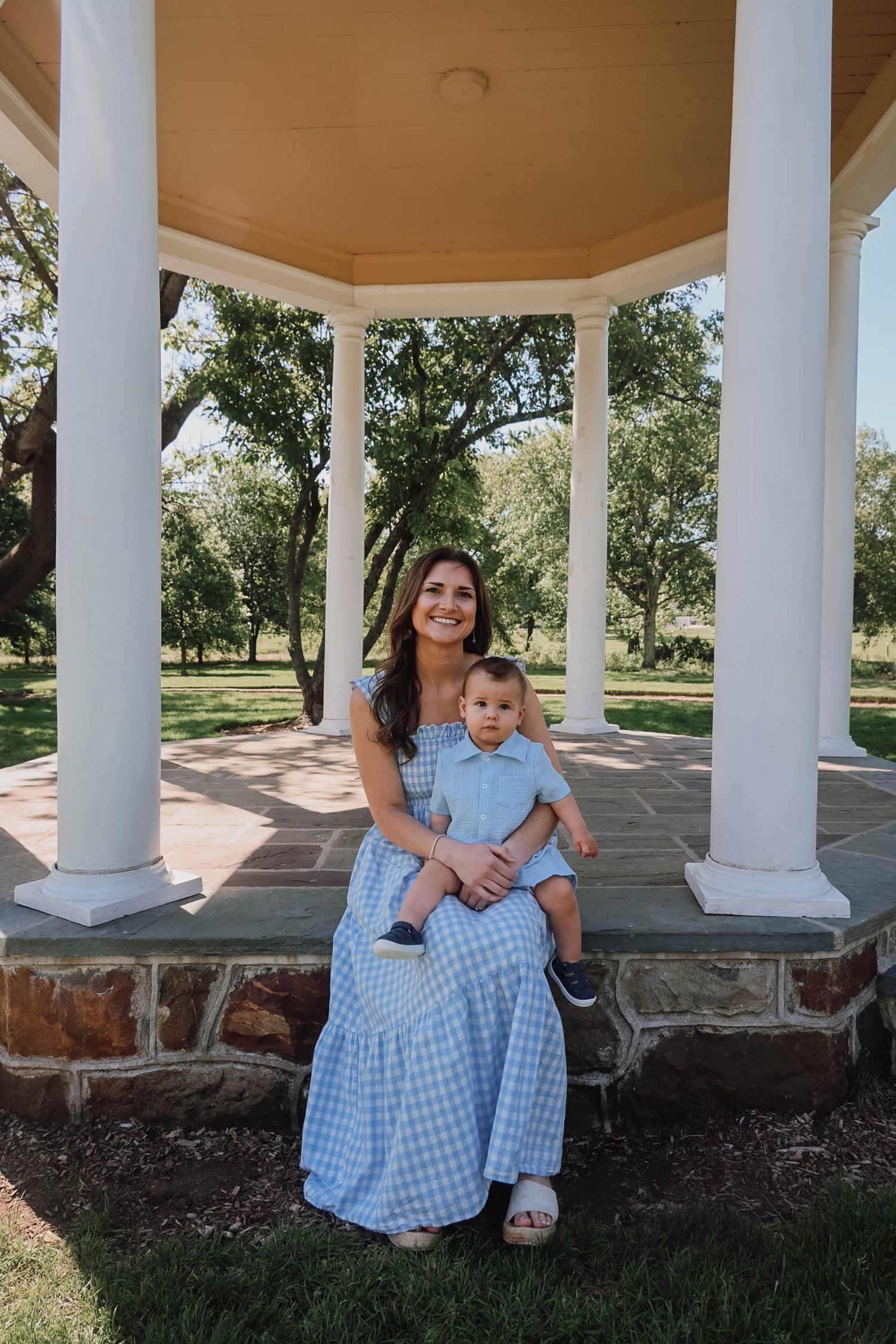 Woman in blue gingham dress sits with baby boy under white gazebo in park.