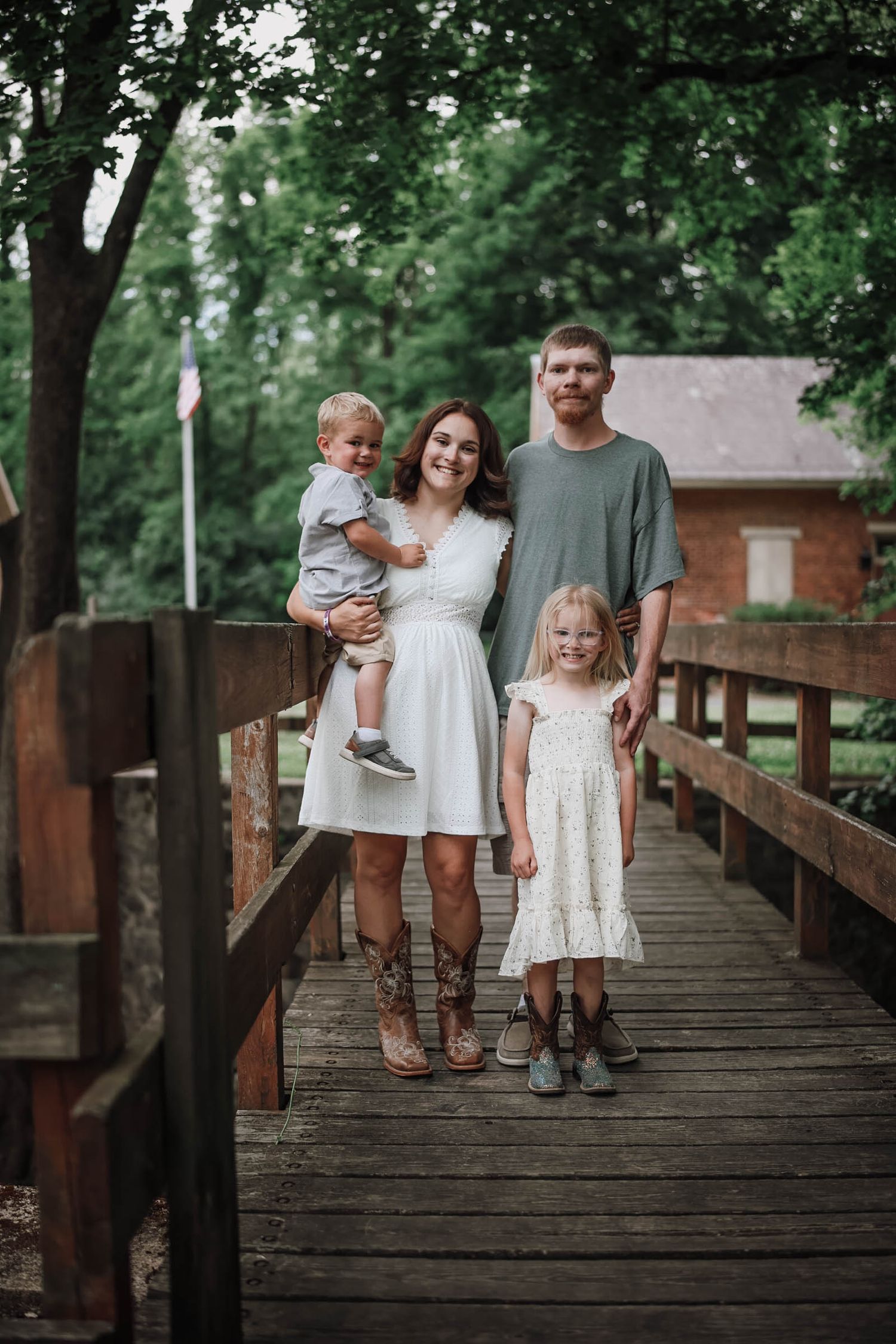 Family of four posing on a wooden bridge surrounded by trees, mom in white dress holding toddler, dad and daughter beside her.
