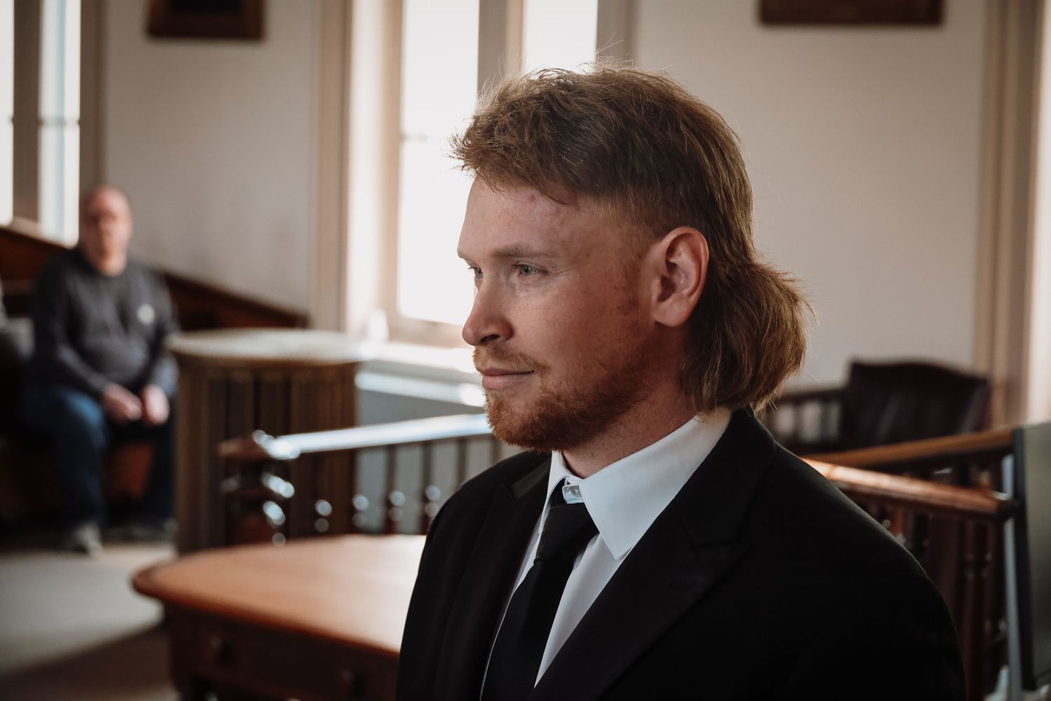 Man with mullet hairstyle and beard wearing black suit and tie standing in a courtroom setting.