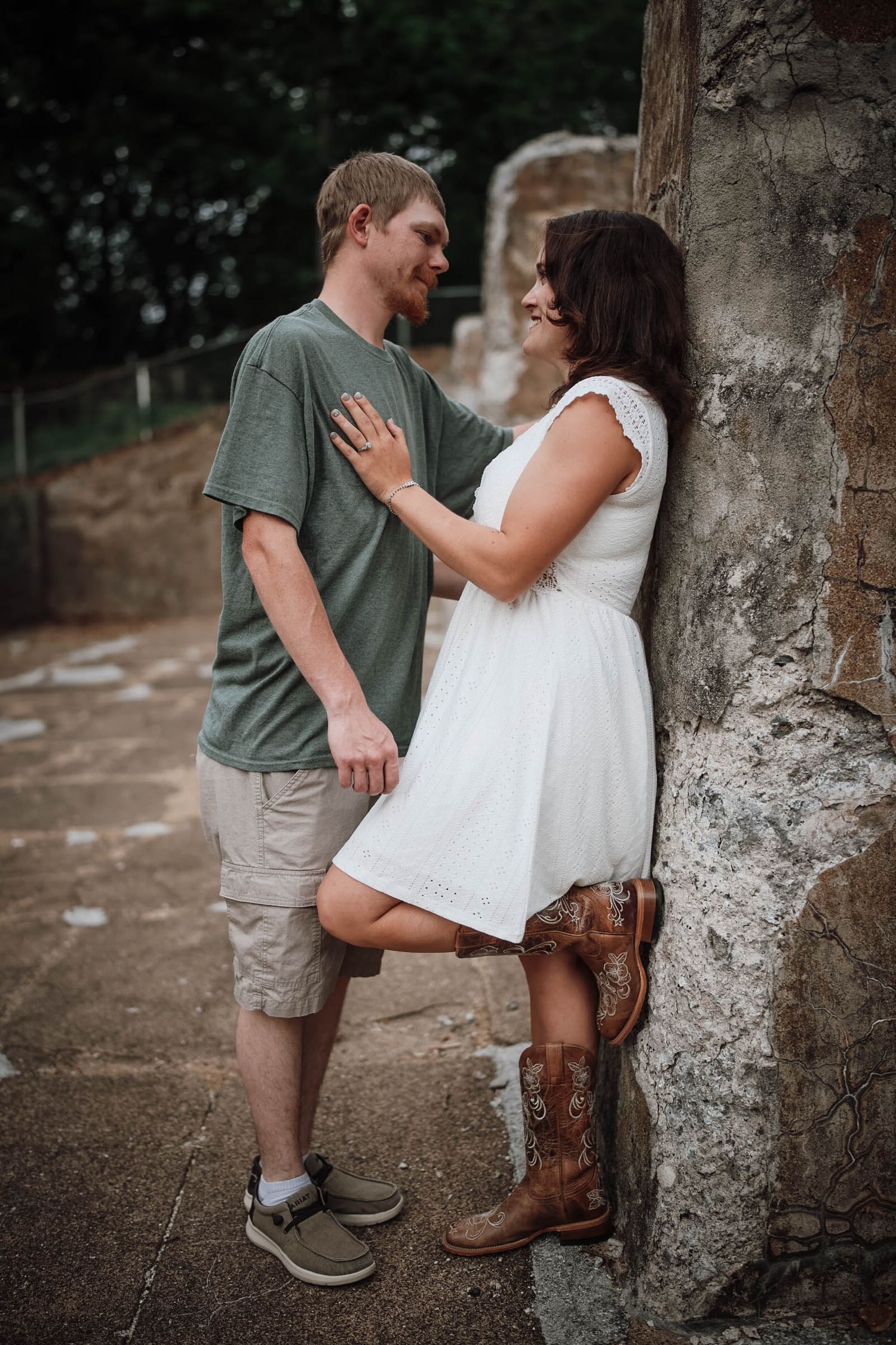 Couple sharing a romantic moment leaning against a stone wall, woman in white dress and cowboy boots.