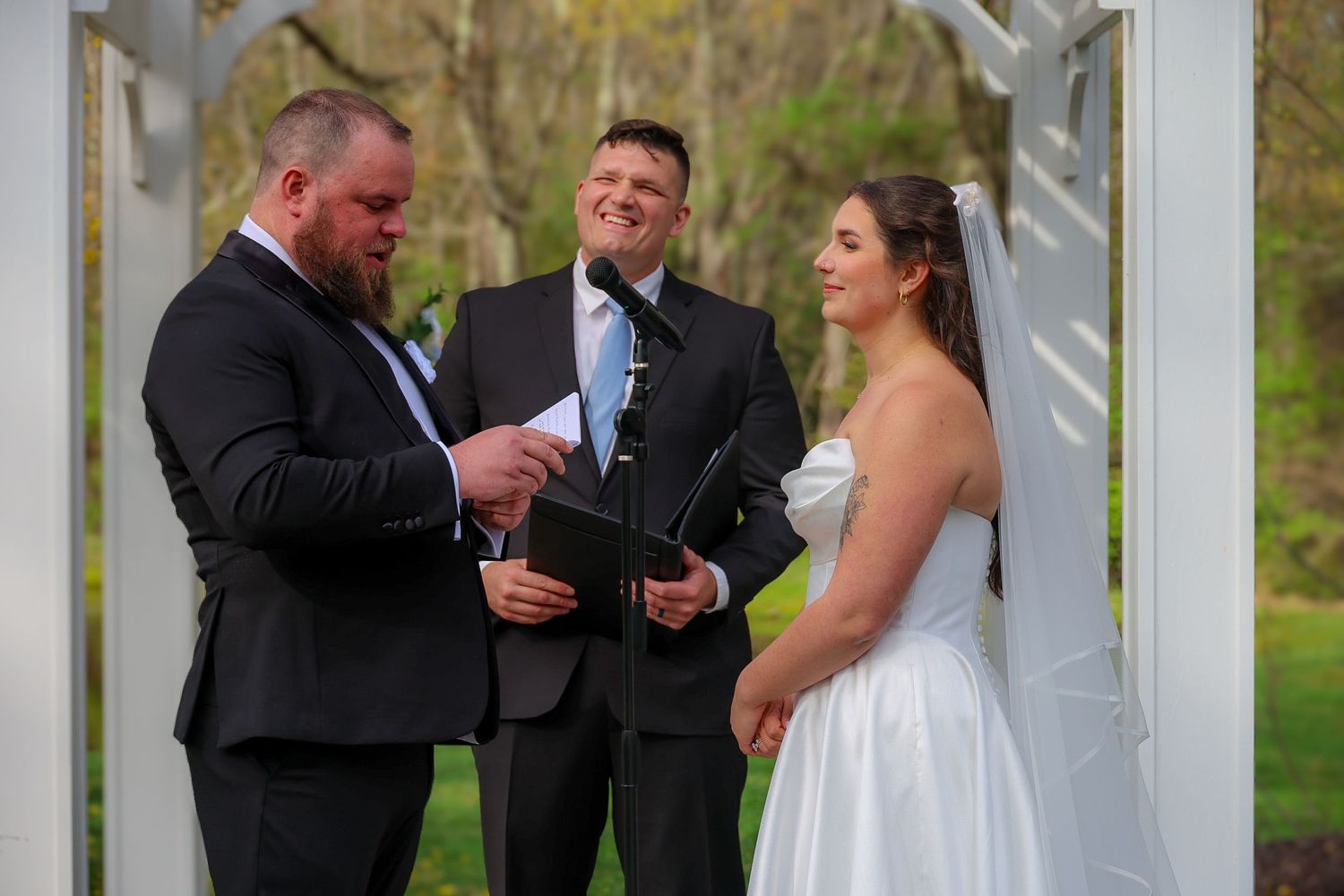 Outdoor wedding ceremony with groom reading vows, smiling officiant, and bride in white gown under white arbor.