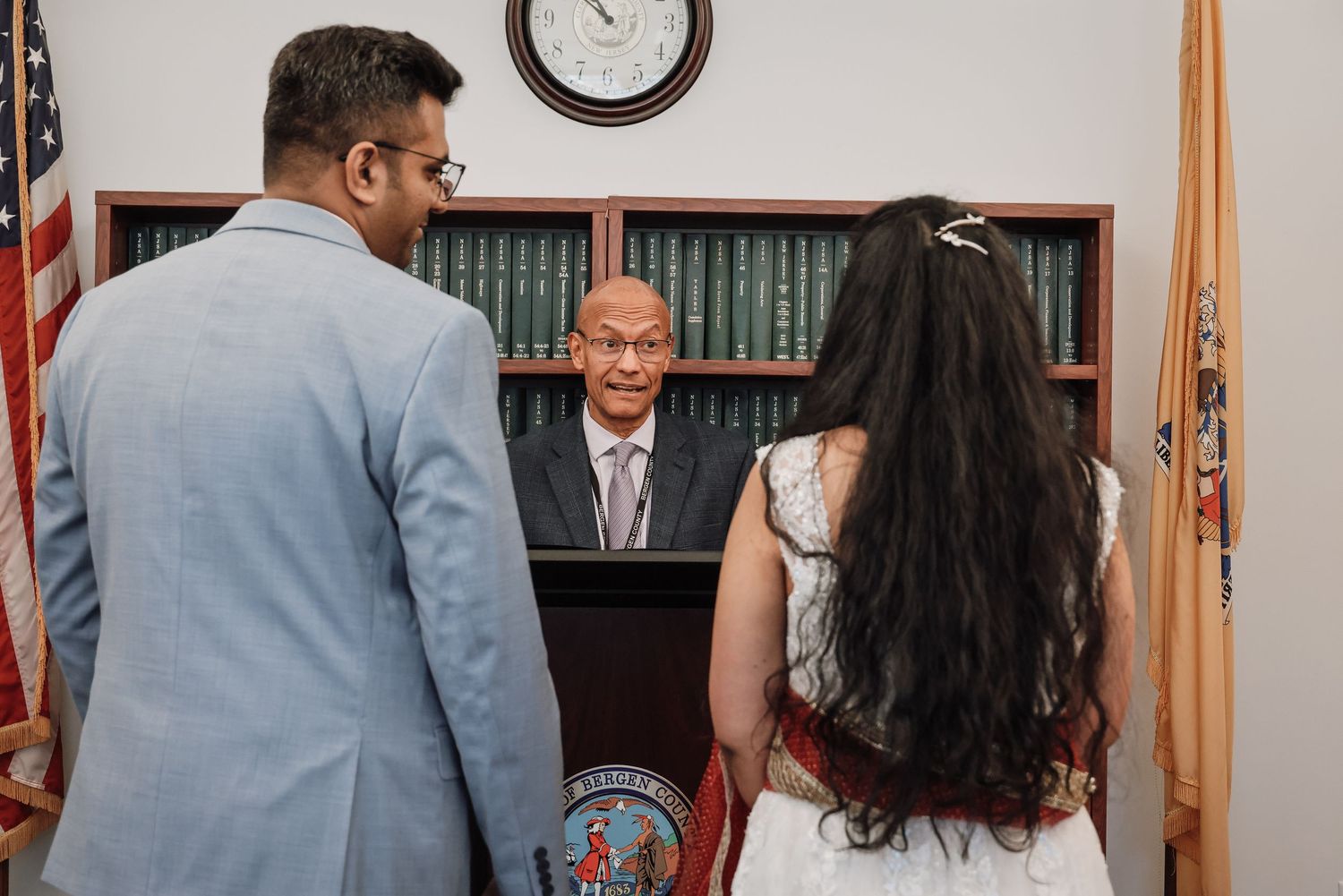 People in formal attire gather in an office setting with an American flag and wall clock visible in the background.
