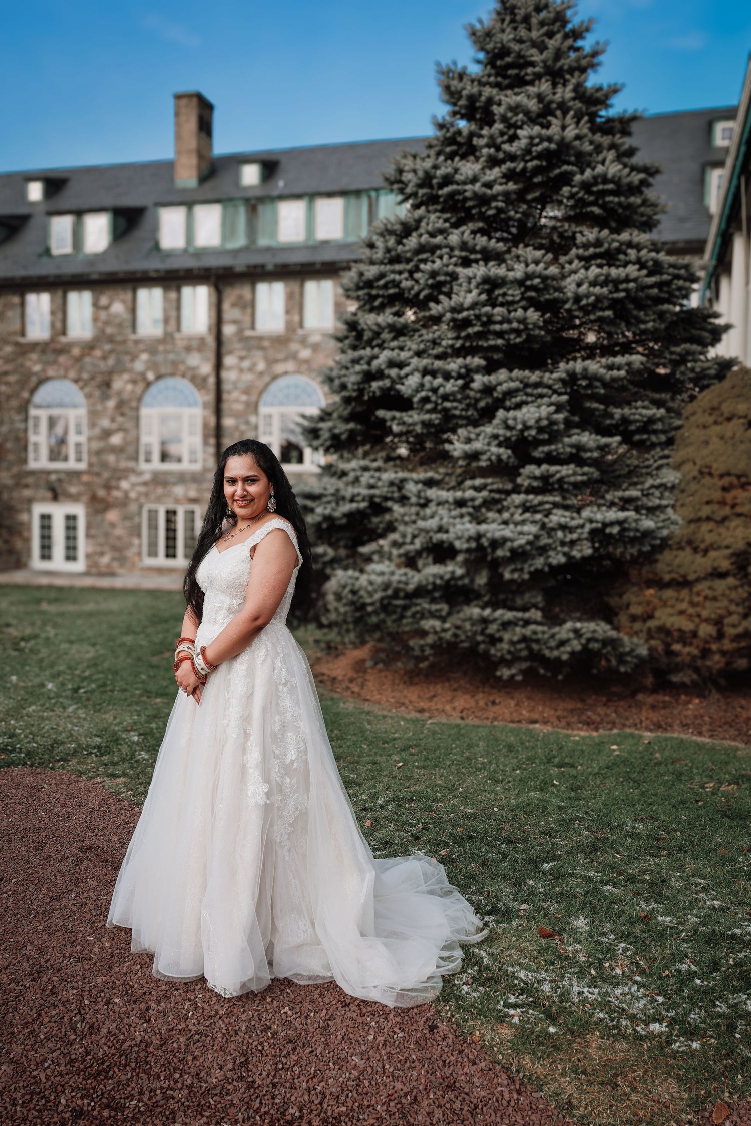 A bride in a flowing white lace dress poses on a lawn with a stone building and evergreen trees in the background.