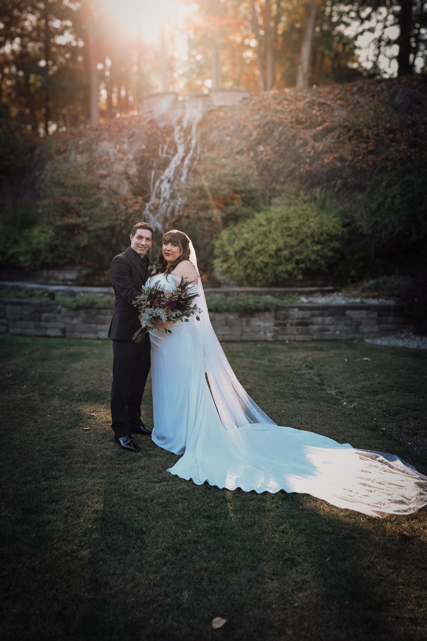 A couple poses for a wedding photo at sunset in front of a stone wall and waterfall in a forest setting.