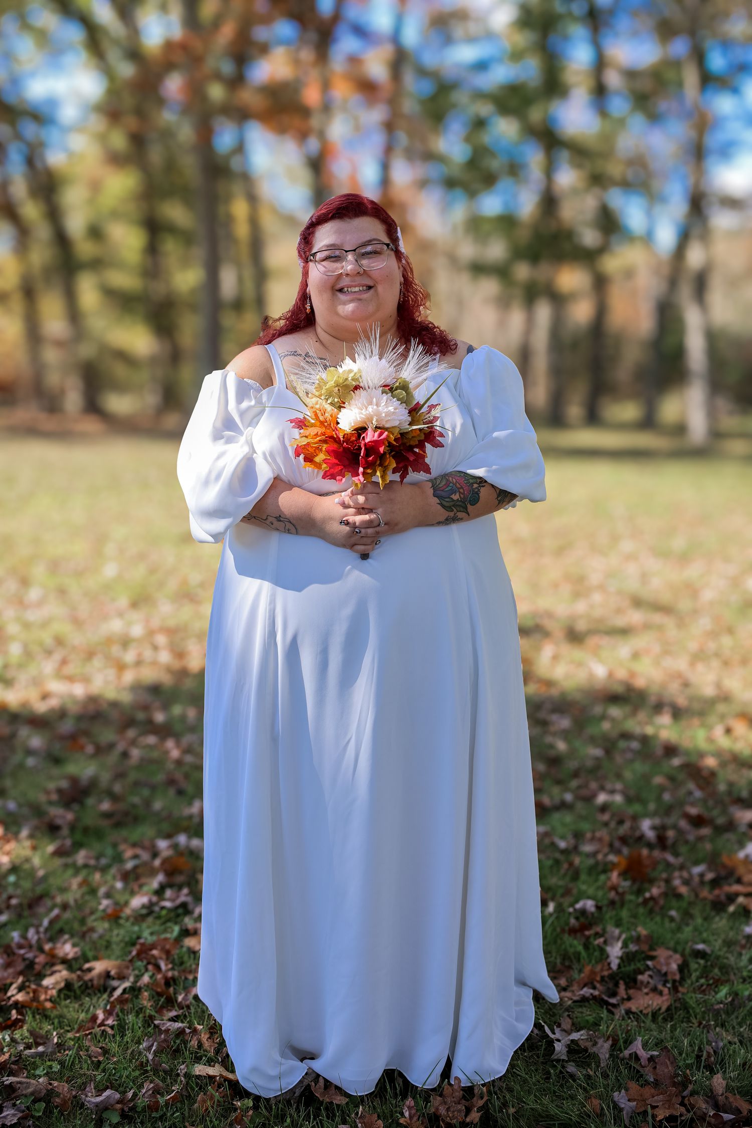 A person in a white dress holds a vibrant autumn bouquet of orange and red flowers in a fall outdoor setting.