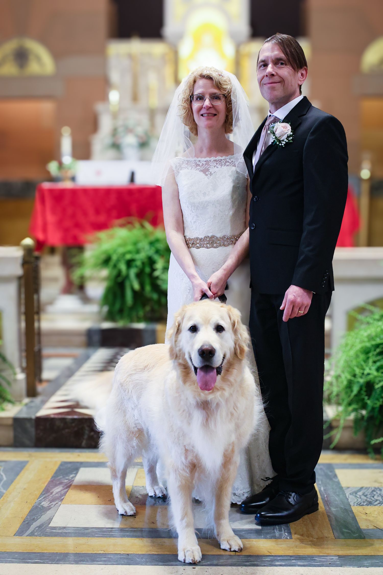 A wedding couple poses with their fluffy white Great Pyrenees dog in a church setting with yellow tile floors.
