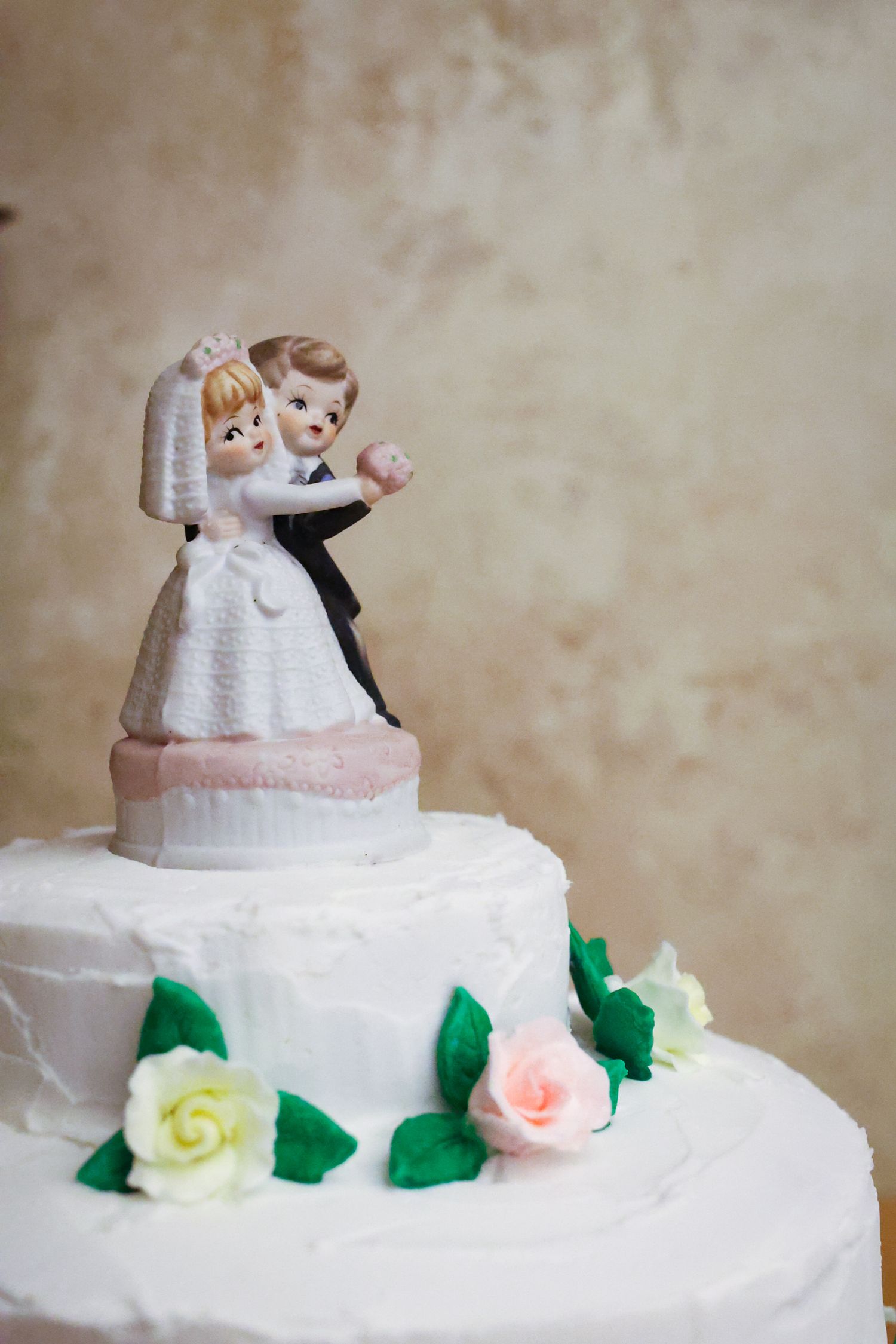 A traditional white wedding cake topper figurine with bride and groom on top of a tiered cake decorated with roses.