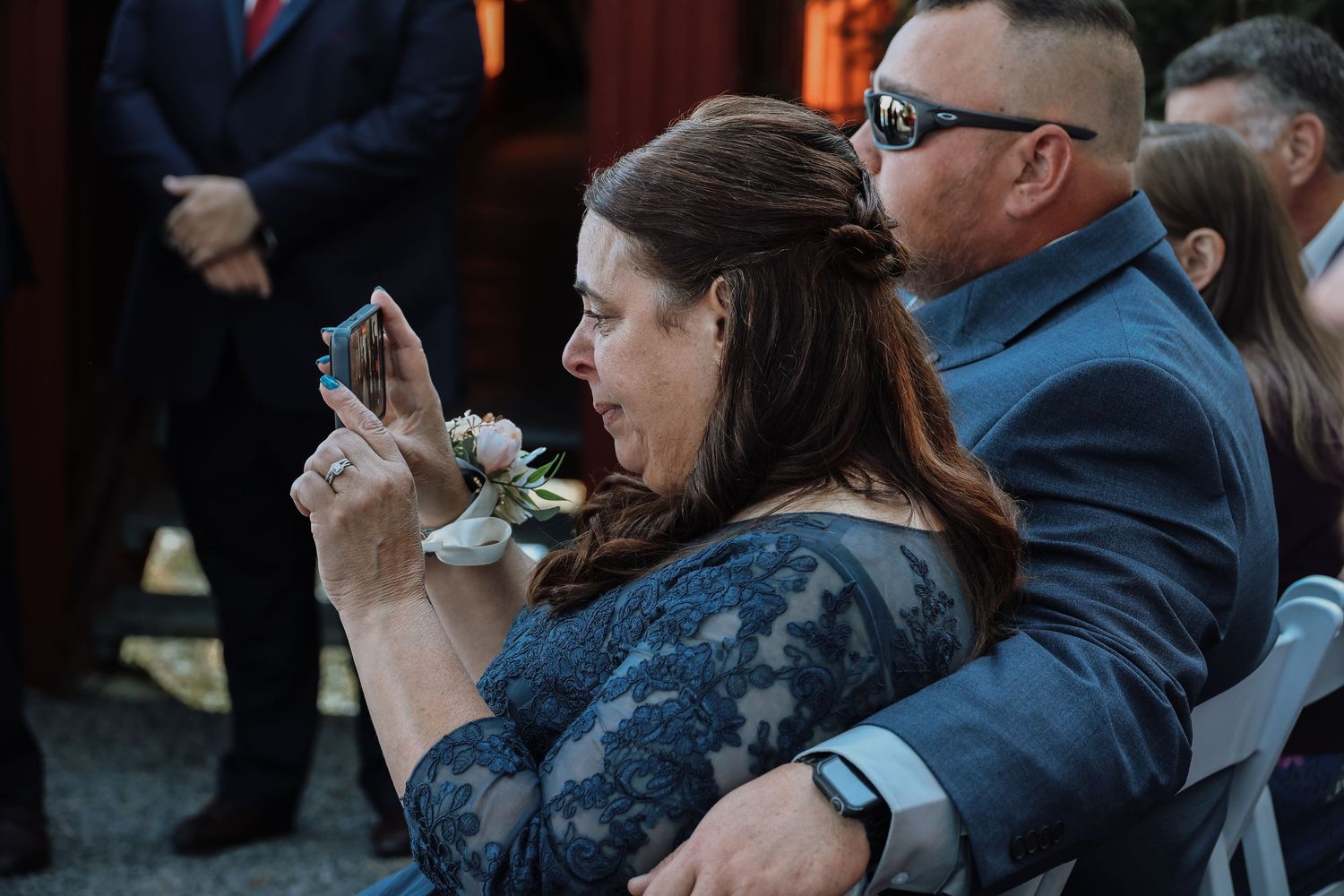 Guest in a navy blue dress takes a photo with their phone during an outdoor event.