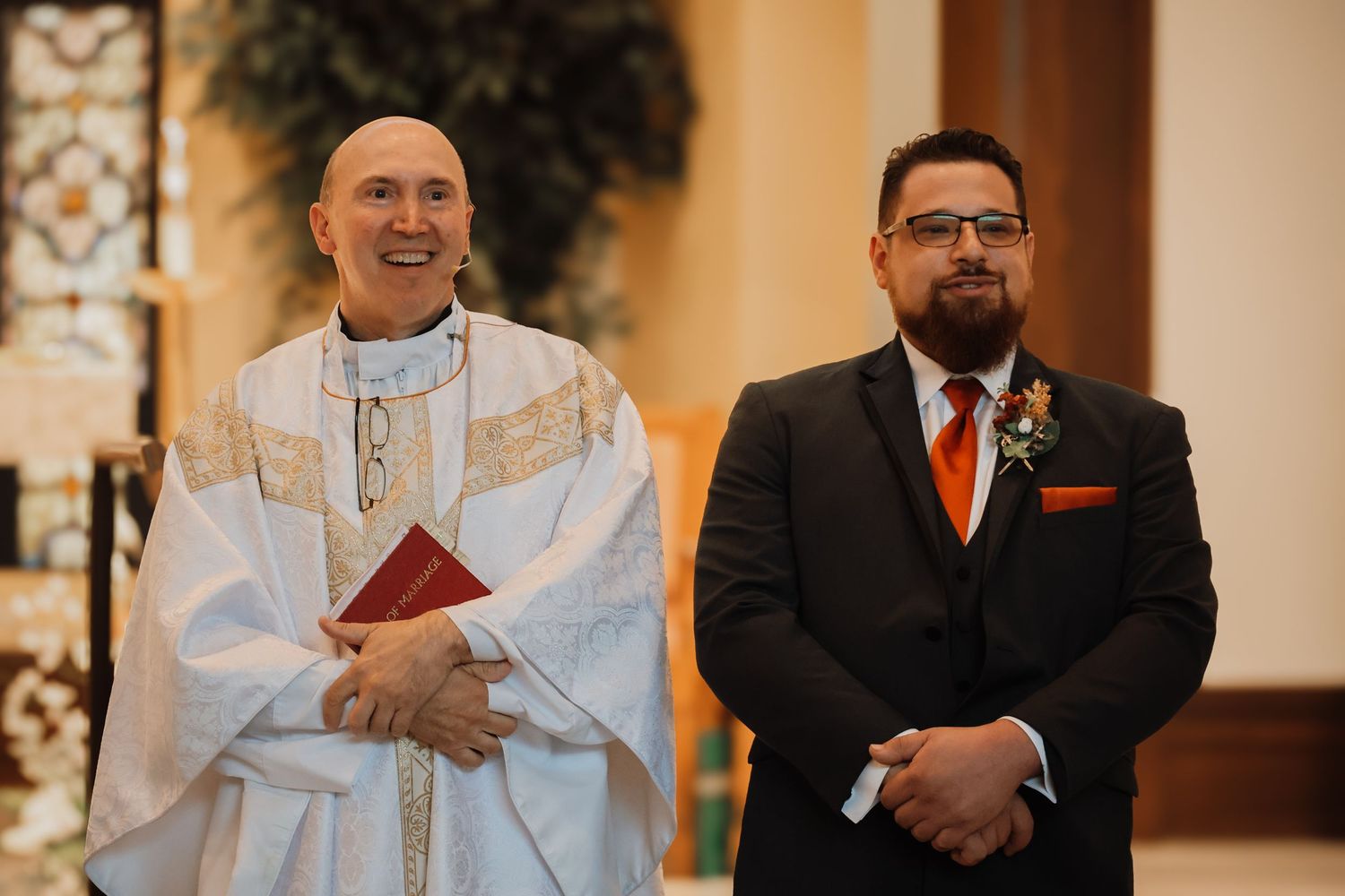 A clergy member in white robes stands next to a groom in a black suit during a wedding ceremony.