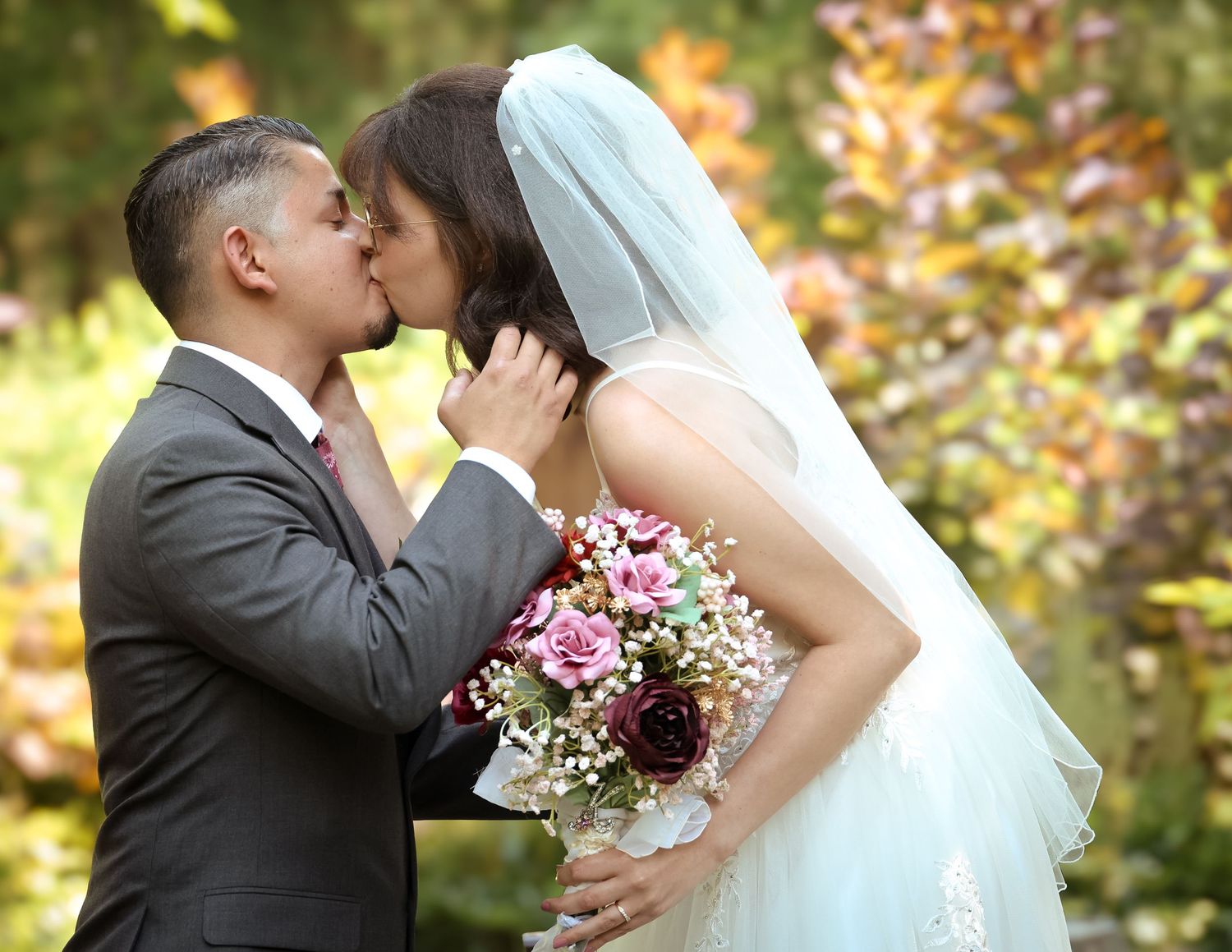 Newlywed couple shares romantic kiss in autumn garden setting with colorful fall foliage in background.