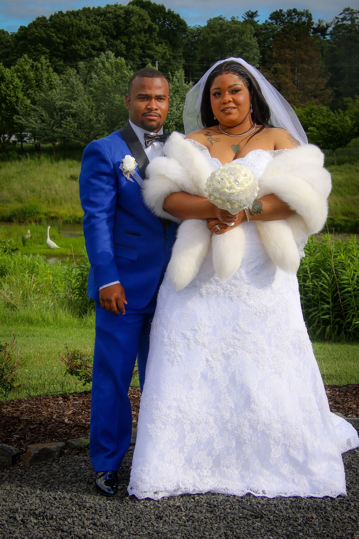 A wedding couple poses outdoors in a blue suit and white lace gown with fur wrap against a green natural backdrop.