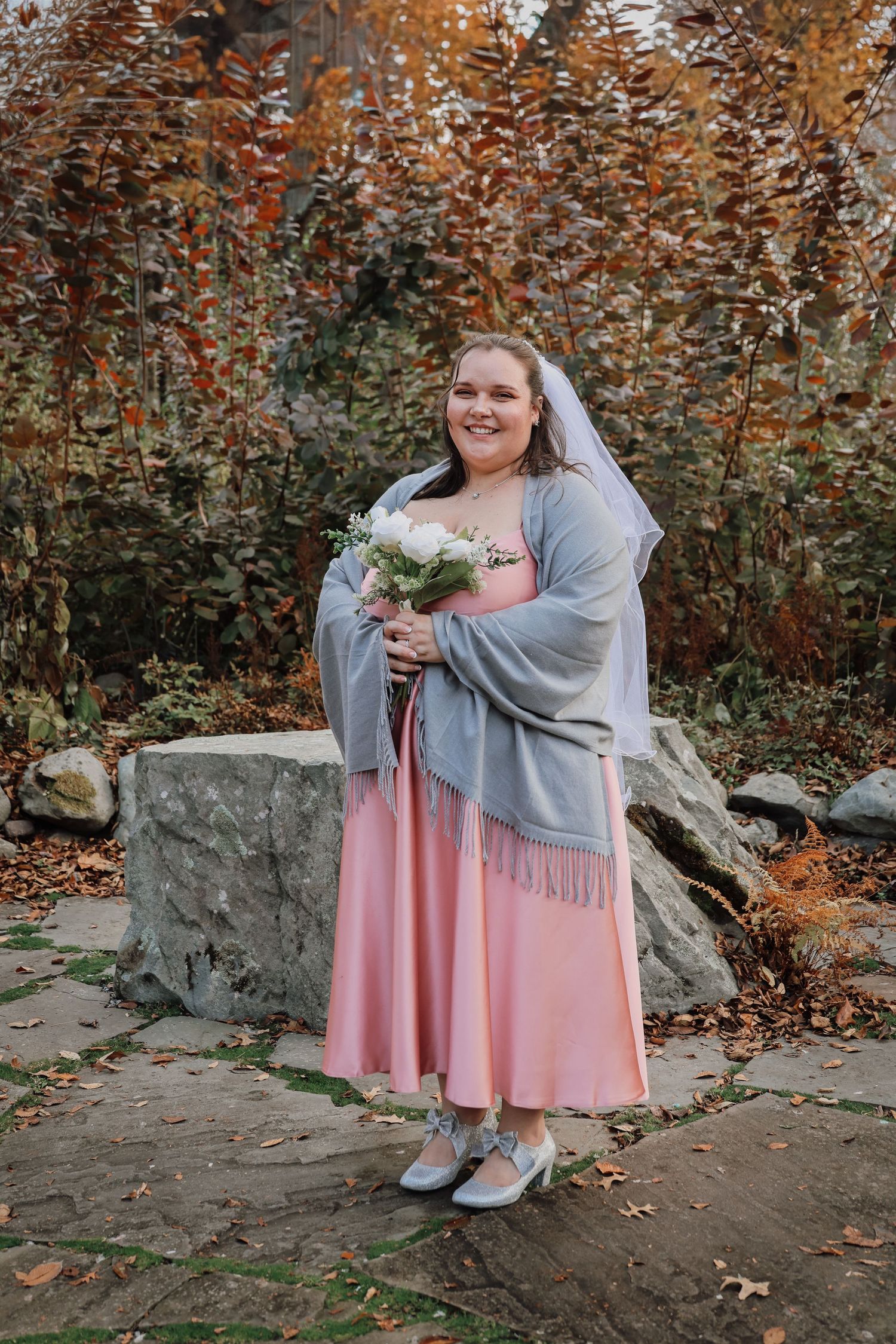 A wedding scene with a pink dress and grey shawl set against autumn foliage and stone walls in a garden setting.