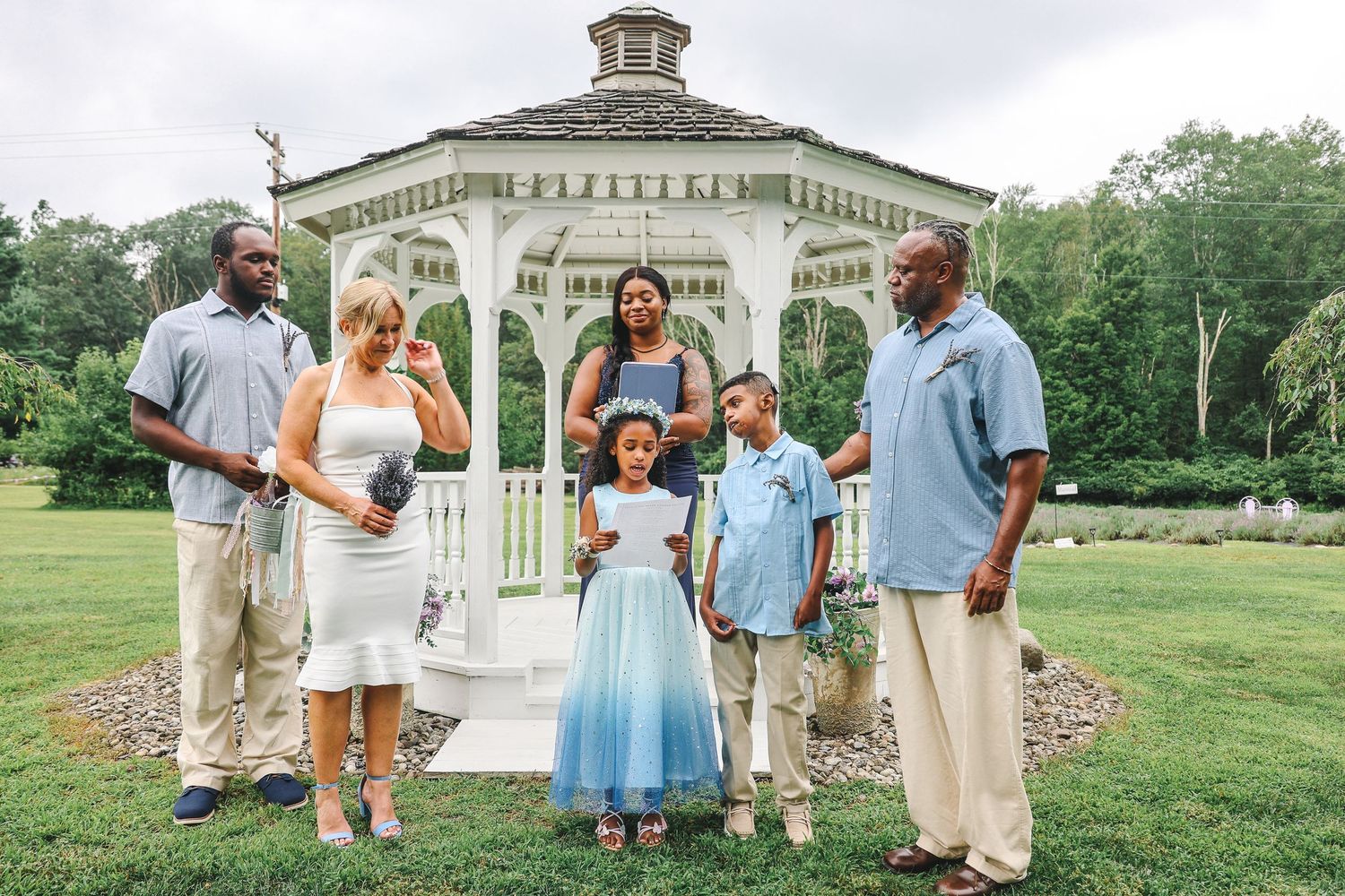 A wedding party gathers in front of a classic white gazebo for outdoor ceremony photos.
