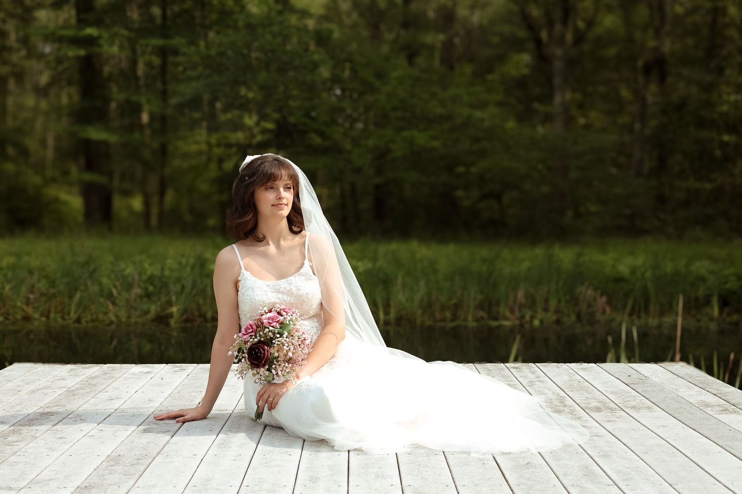A bride in a white wedding dress sits gracefully on a wooden dock surrounded by lush green trees and nature.