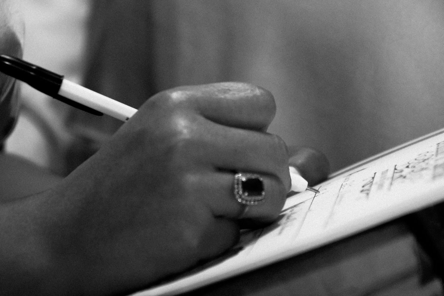 A person's hand writes with a pen on paper in black and white, creating a dramatic close-up composition.