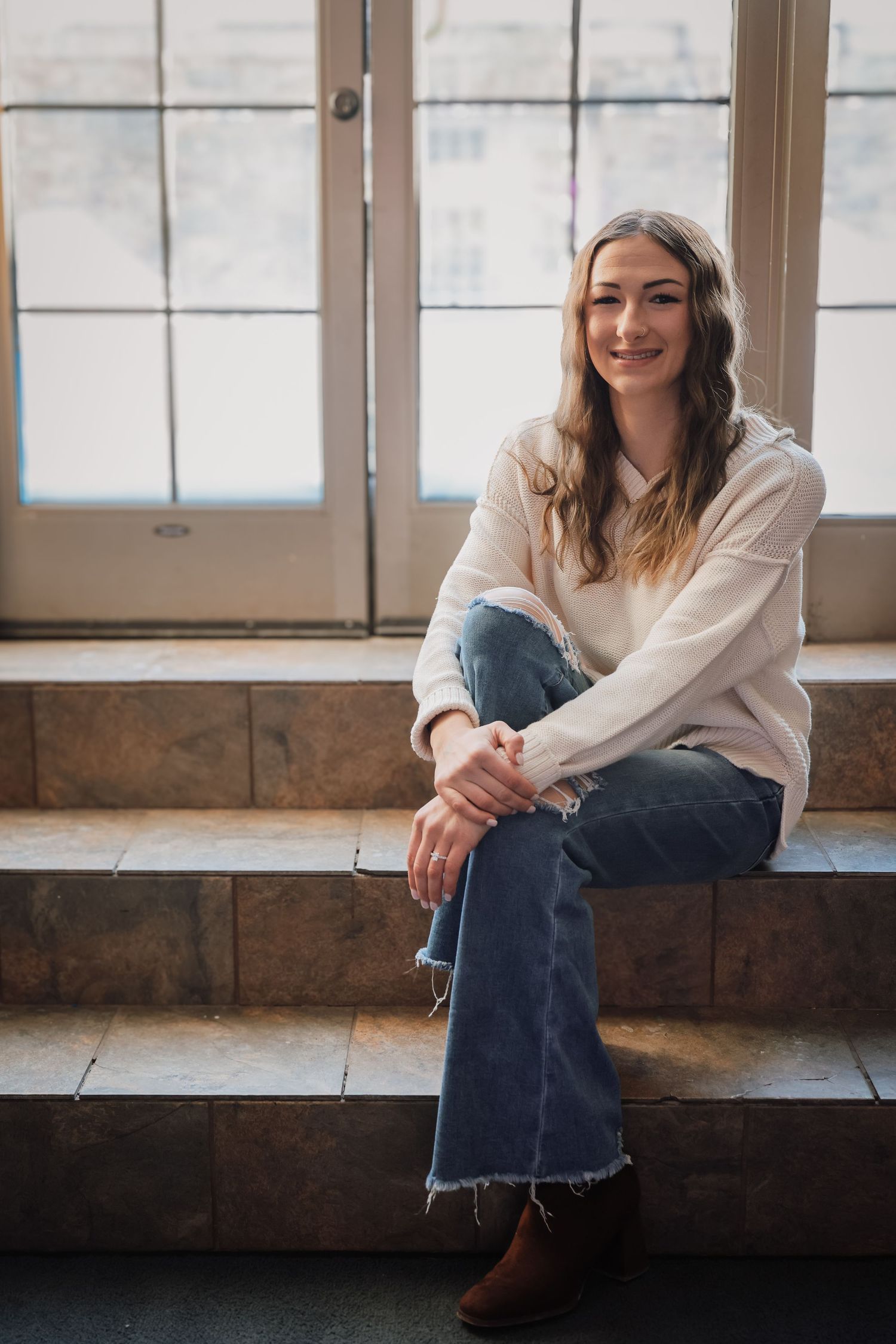 A person in a white sweater and jeans sits casually on stone steps near French doors in natural lighting.