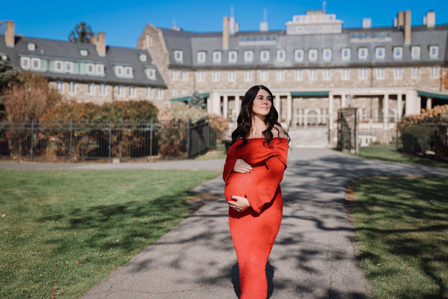 A person in a bright red coat stands on a gravel path in front of a historic stone building on a sunny day.