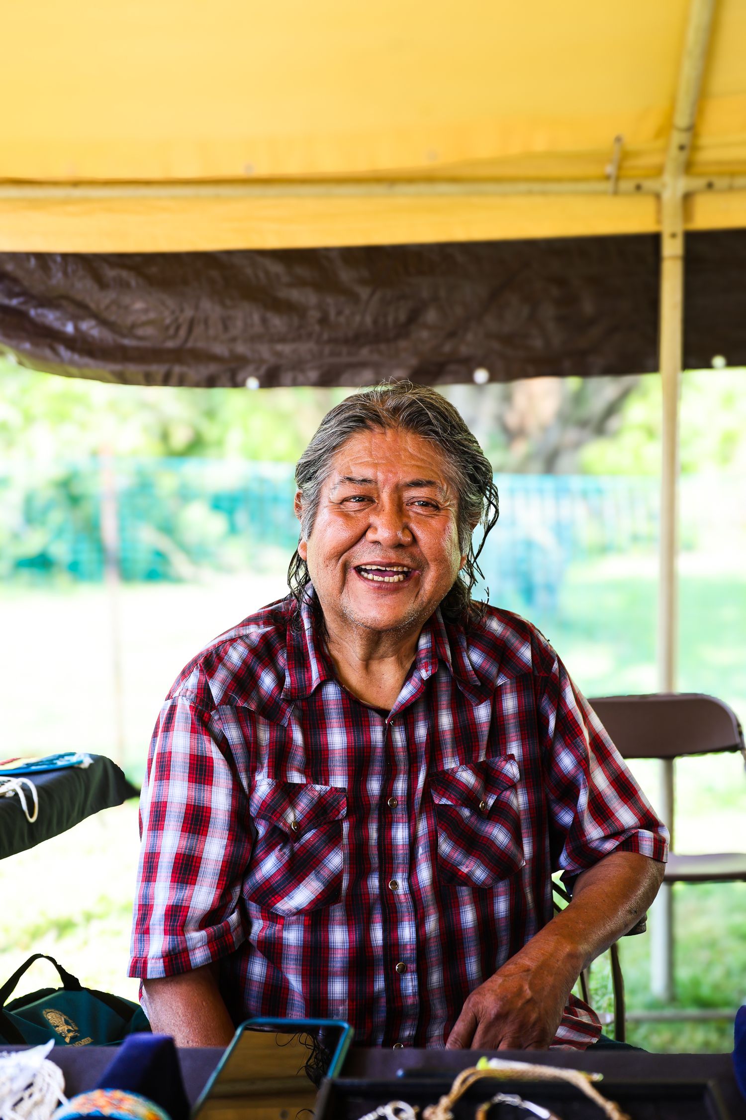 A person in a plaid shirt sits outdoors under a covered area and smiles warmly at the camera.