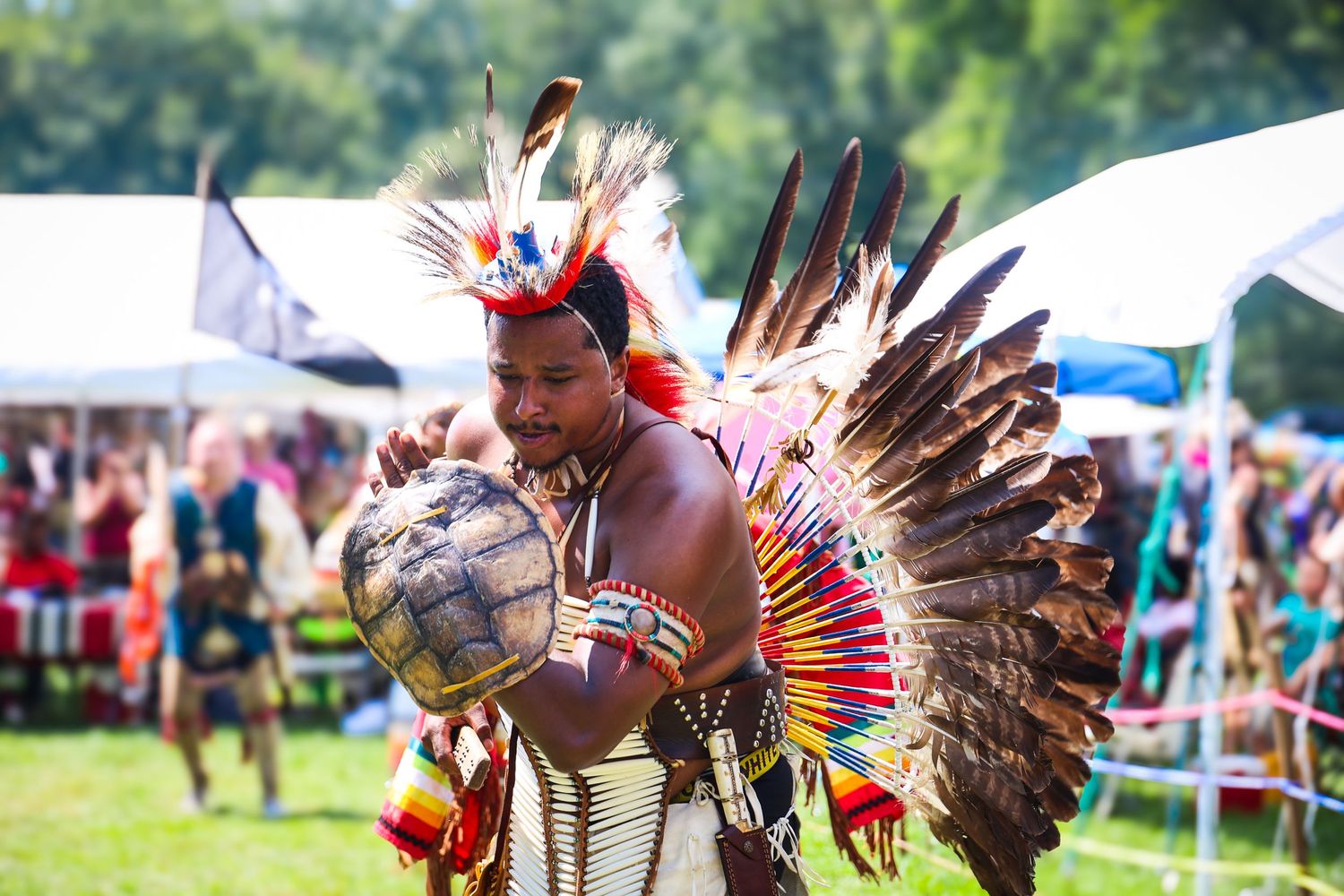 Native American dancers in traditional regalia perform at an outdoor powwow festival with colorful feathers and outfits.
