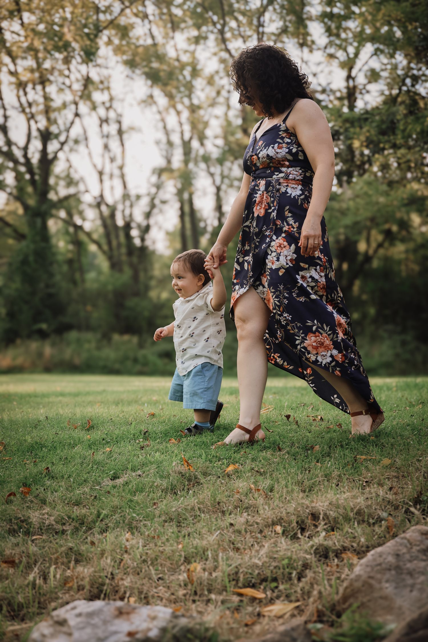 A woman in a floral dress walks with a small child in a t-shirt and shorts on grassy ground during golden hour.