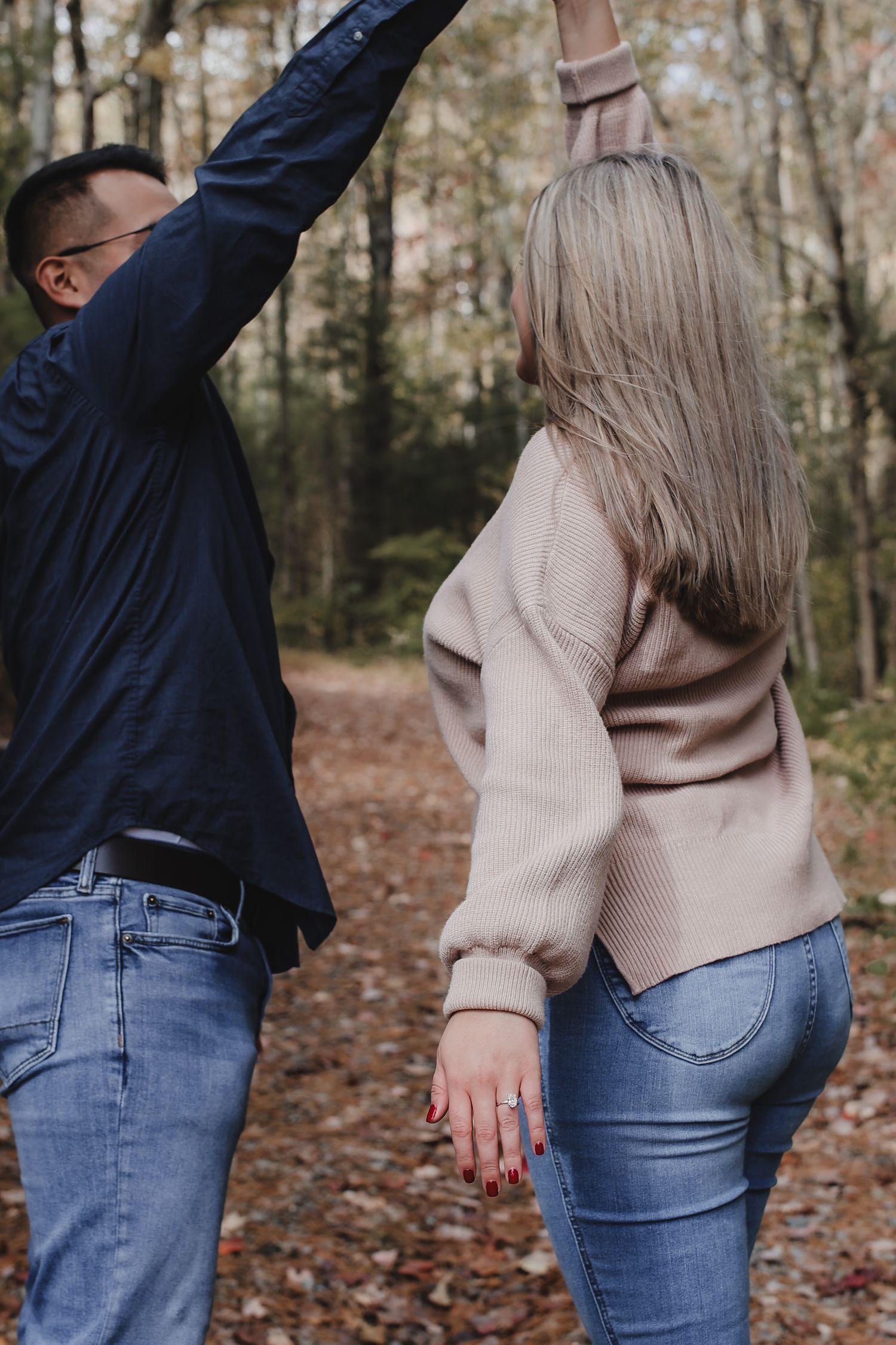 A couple in jeans and sweaters poses playfully together in a wooded autumn setting with fallen leaves on the ground.