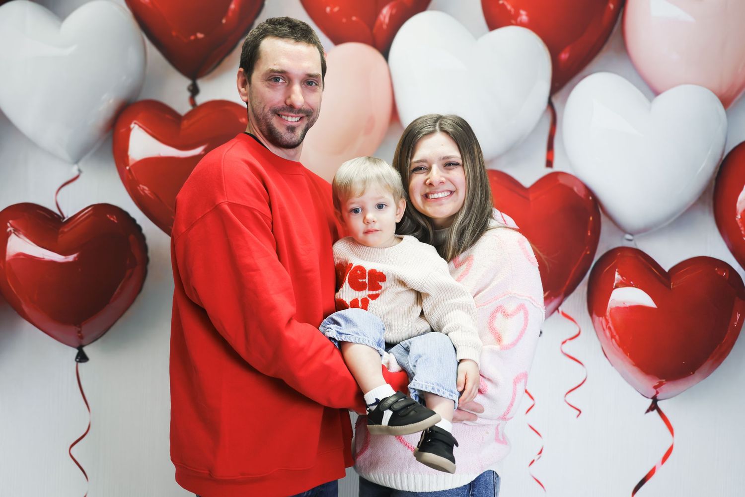 Family wearing red sweaters poses against white and red heart-shaped balloon backdrop for Valentine's Day photos.