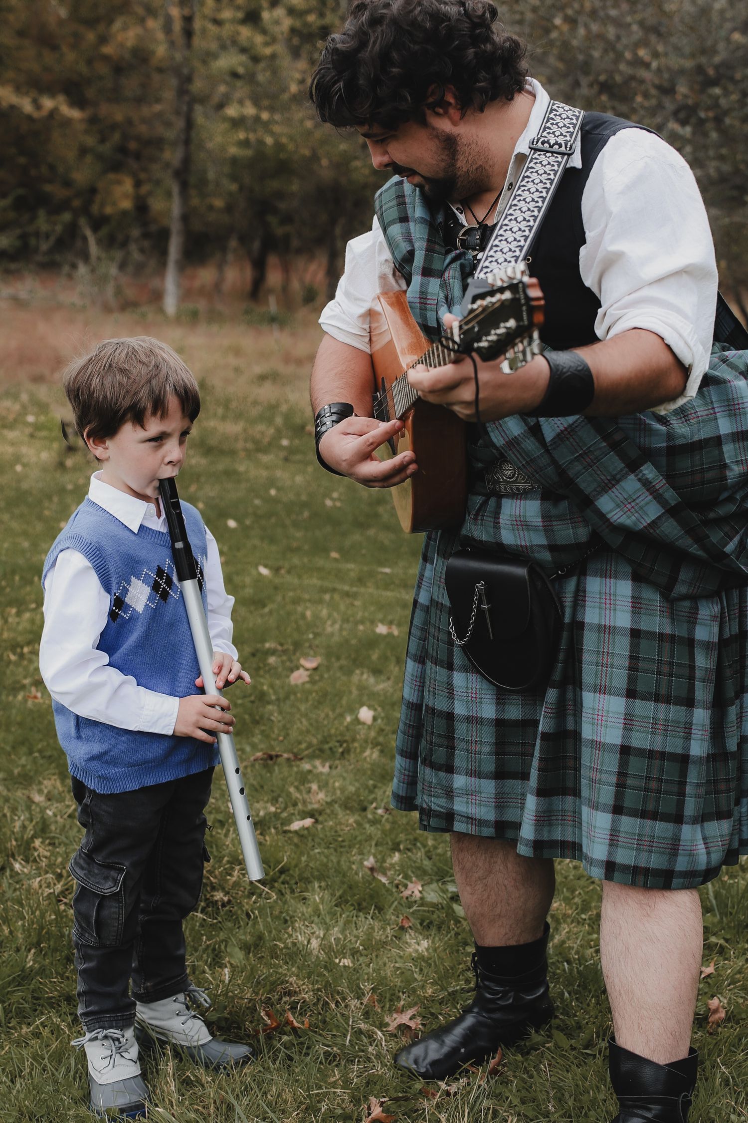 A musician in traditional Scottish kilt plays guitar while a young boy in blue vest holds a recorder in an outdoor setting.
