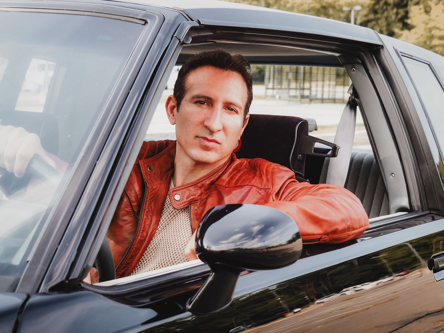 A person in an orange jacket leans out of a car window while driving through a wooded area.