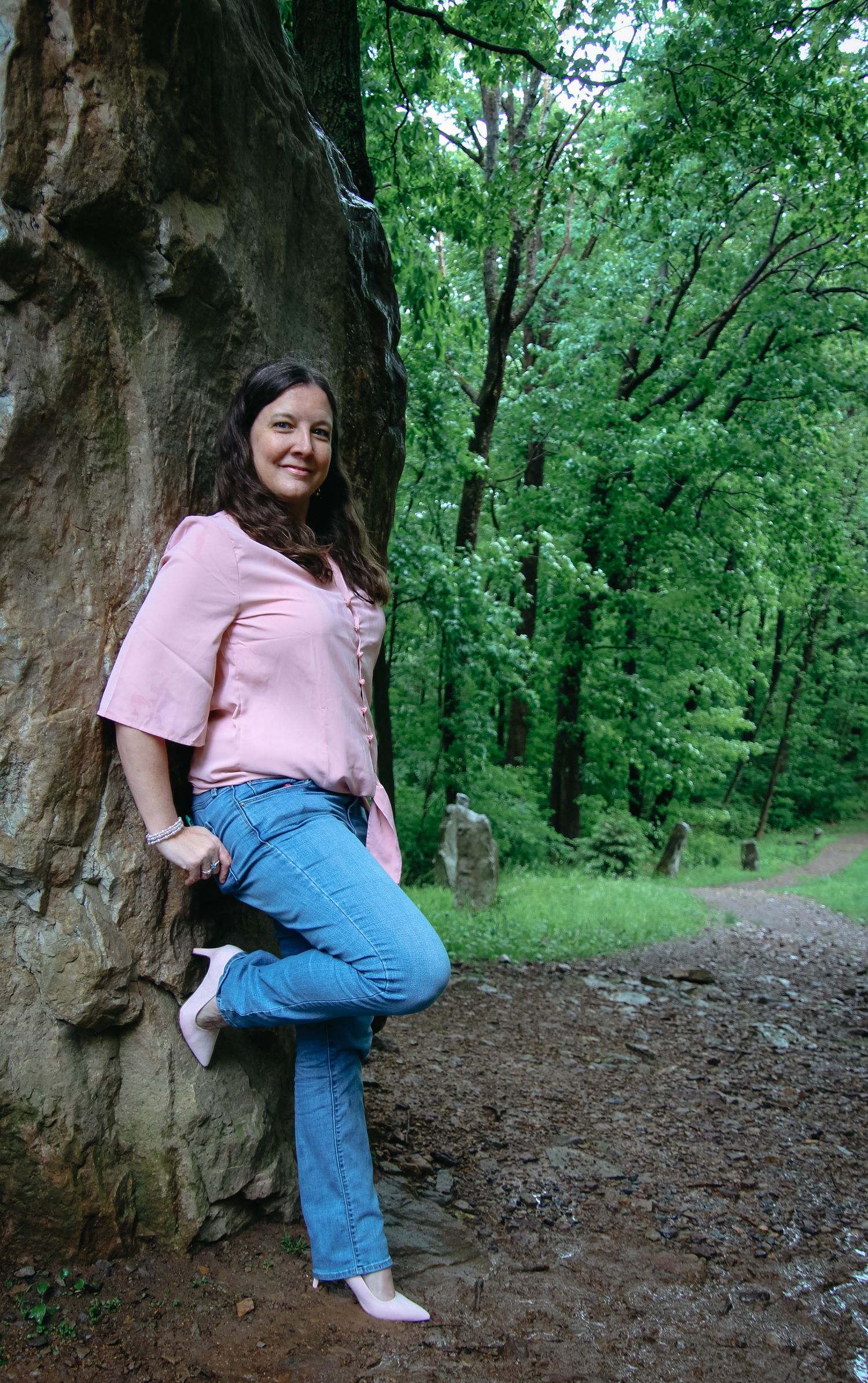 A person in a pink shirt and jeans poses on a rocky outcrop along a wooded trail on a summer day.