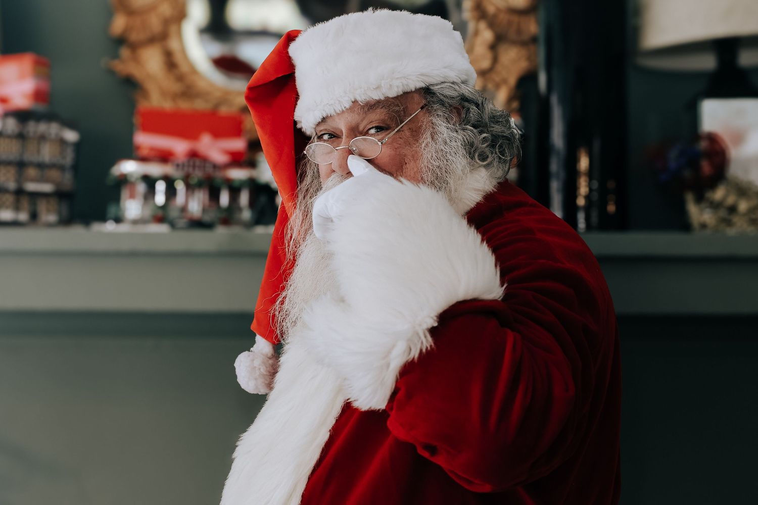 A traditional figure in a red suit and white beard wearing a Santa hat adjusts their glasses indoors.