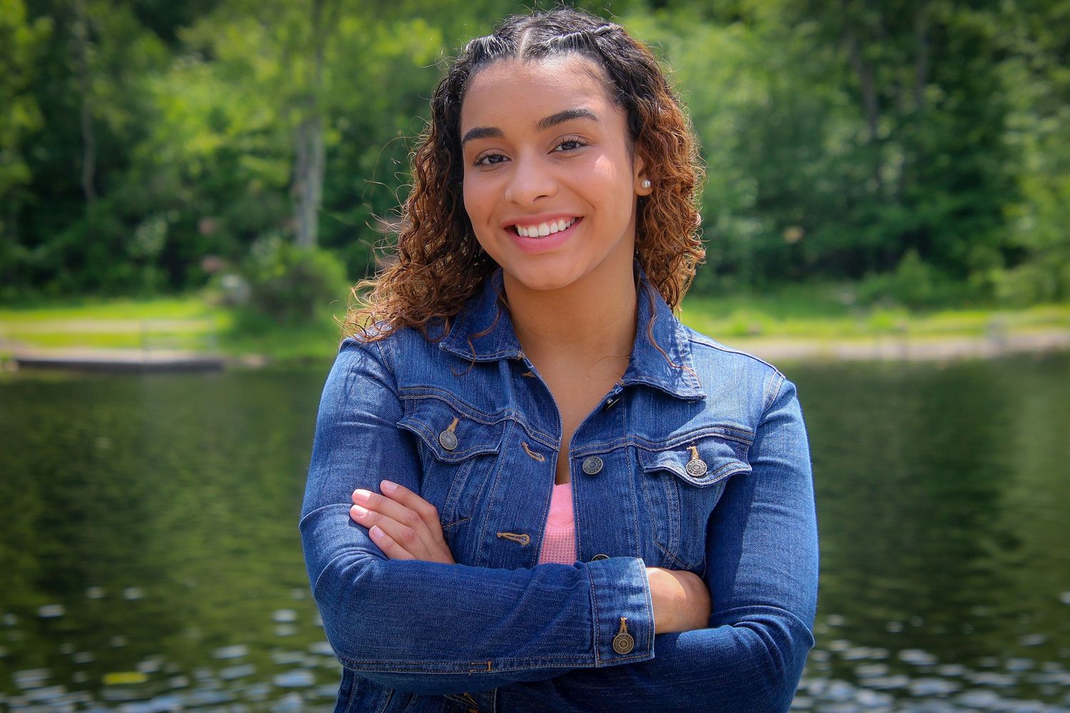 Someone wearing a denim jacket poses outdoors near a scenic lake surrounded by green trees and nature.