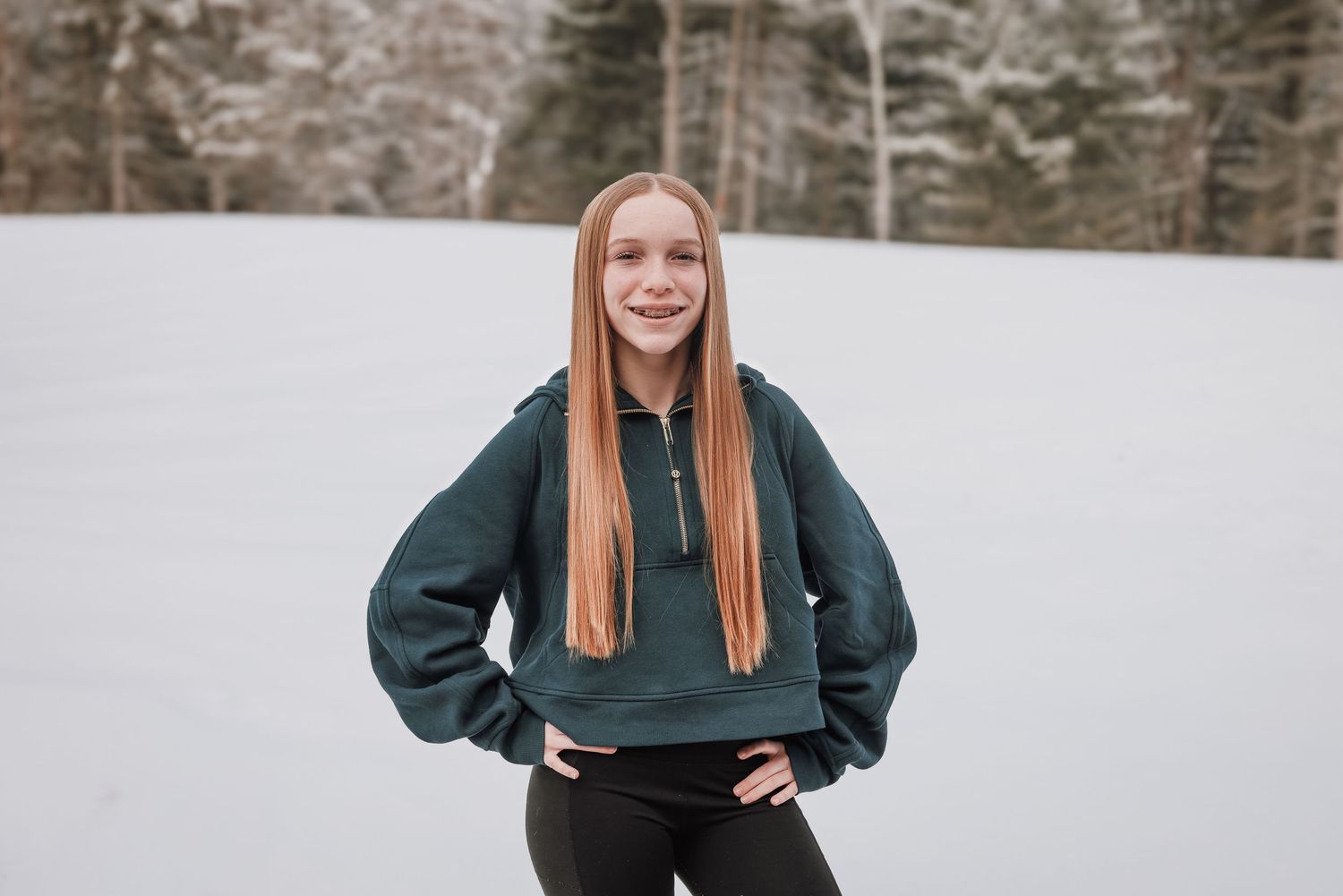 A person with long straight red hair wearing a dark green sweatshirt and black pants stands in front of a snowy background.