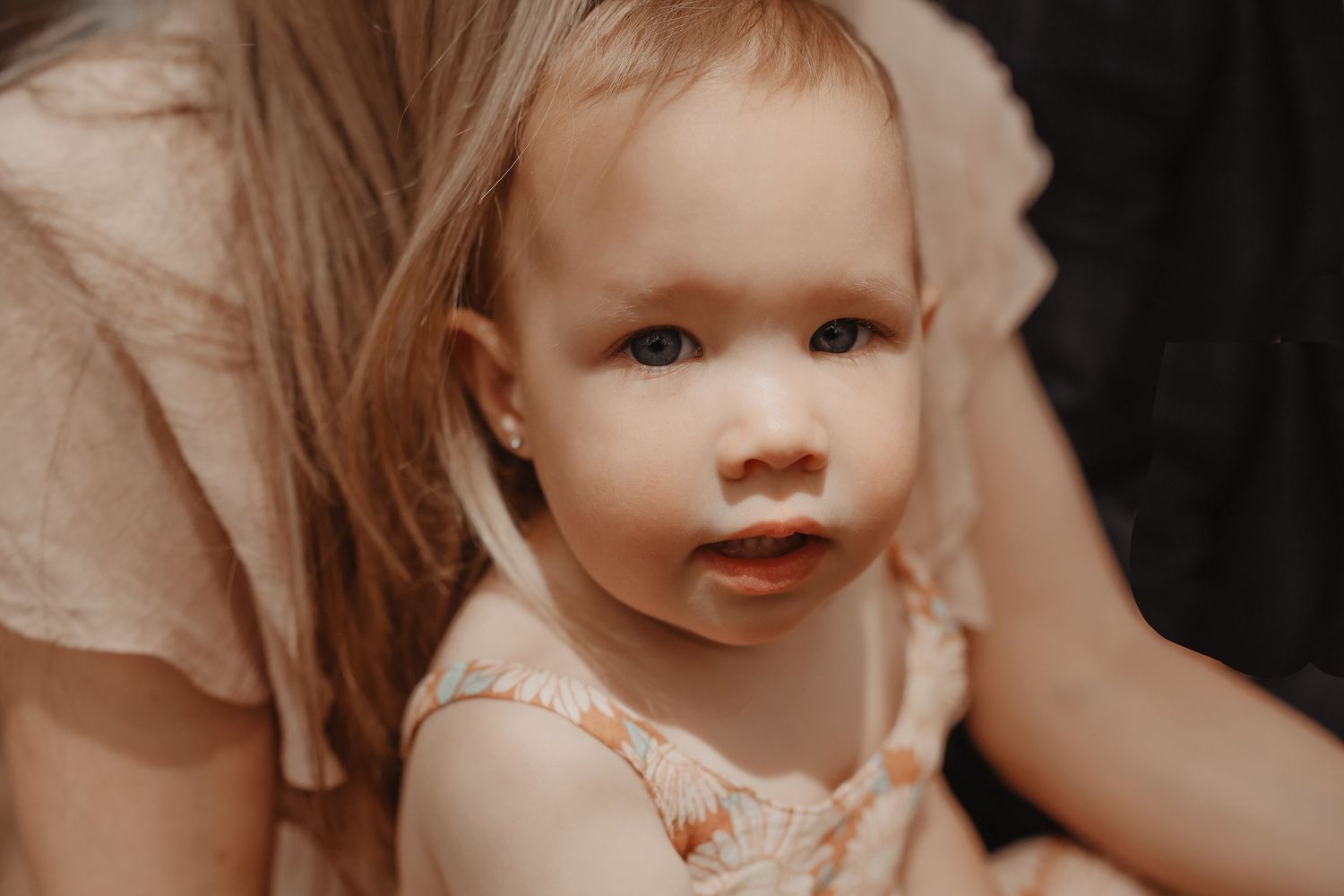 A baby wearing a white dress smiles while being held by someone in a dark setting.