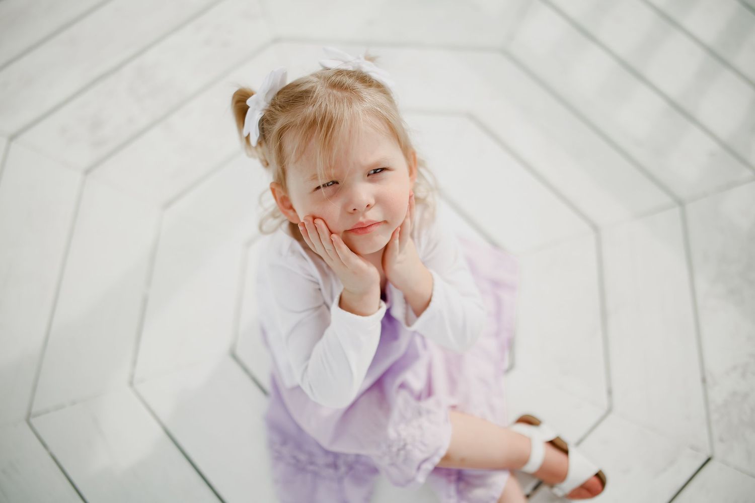 A child in a lavender tulle dress and white cardigan poses playfully on a white wooden floor.