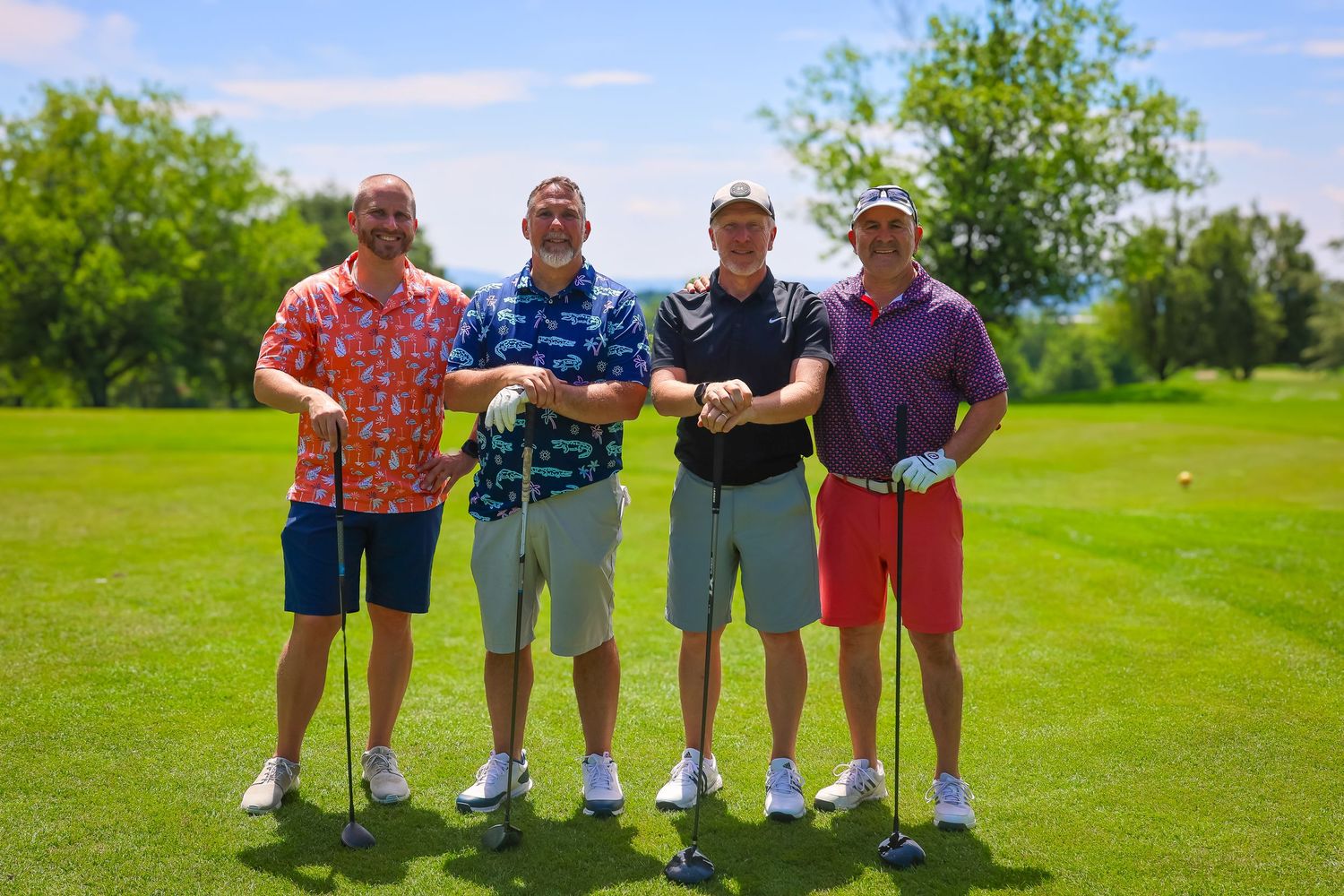 Four golfers stand together with their clubs on a sunny golf course with green grass and trees in the background.