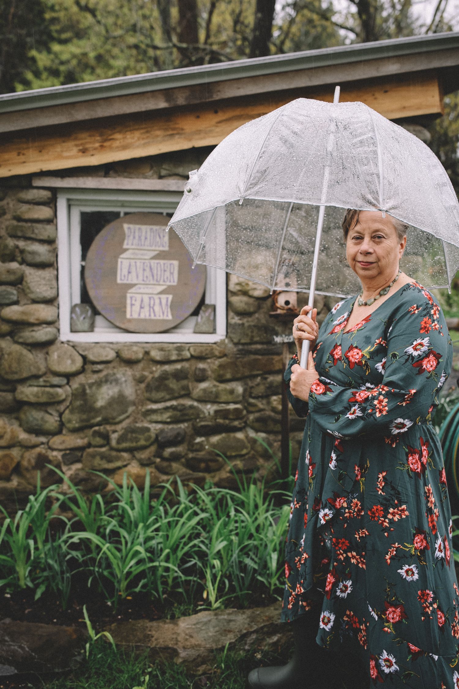 A person in a floral dress holds a clear umbrella while standing against a stone wall in a garden setting.