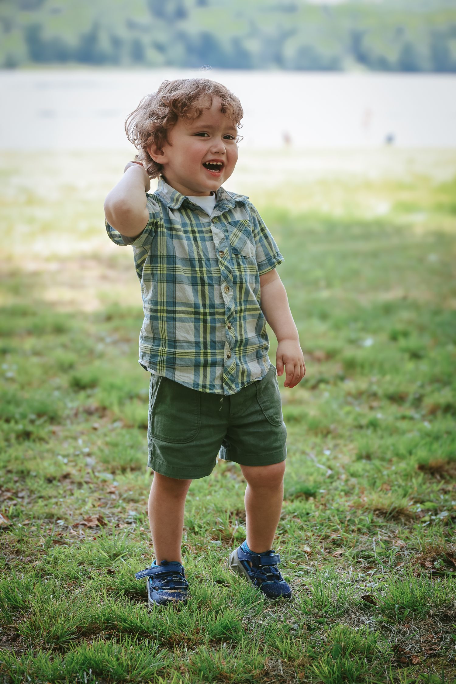 A toddler wearing a plaid shirt and green shorts plays joyfully in a grassy outdoor setting.