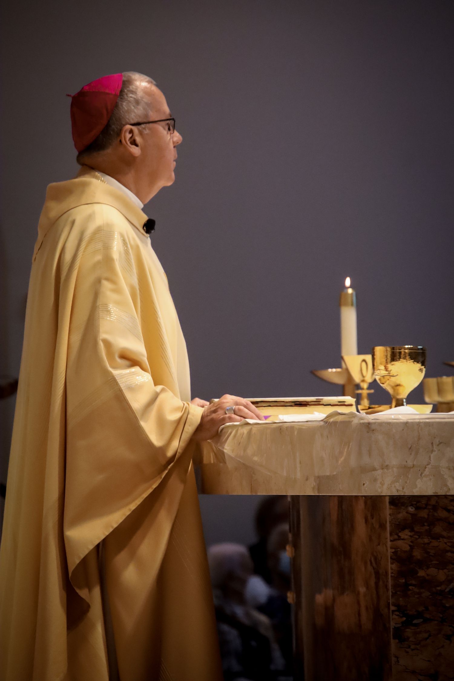 A figure in cream-colored vestments and red cap stands at an altar with lit candle and chalice in a dark church setting.