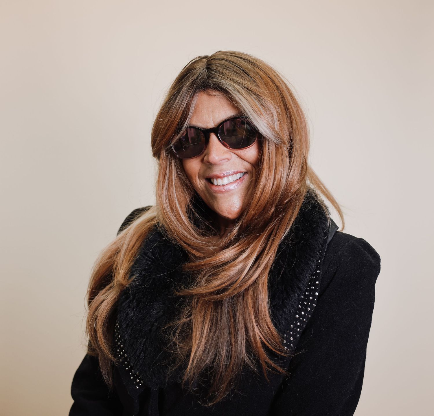Woman with long brown hair wearing sunglasses and black top smiles against neutral background.