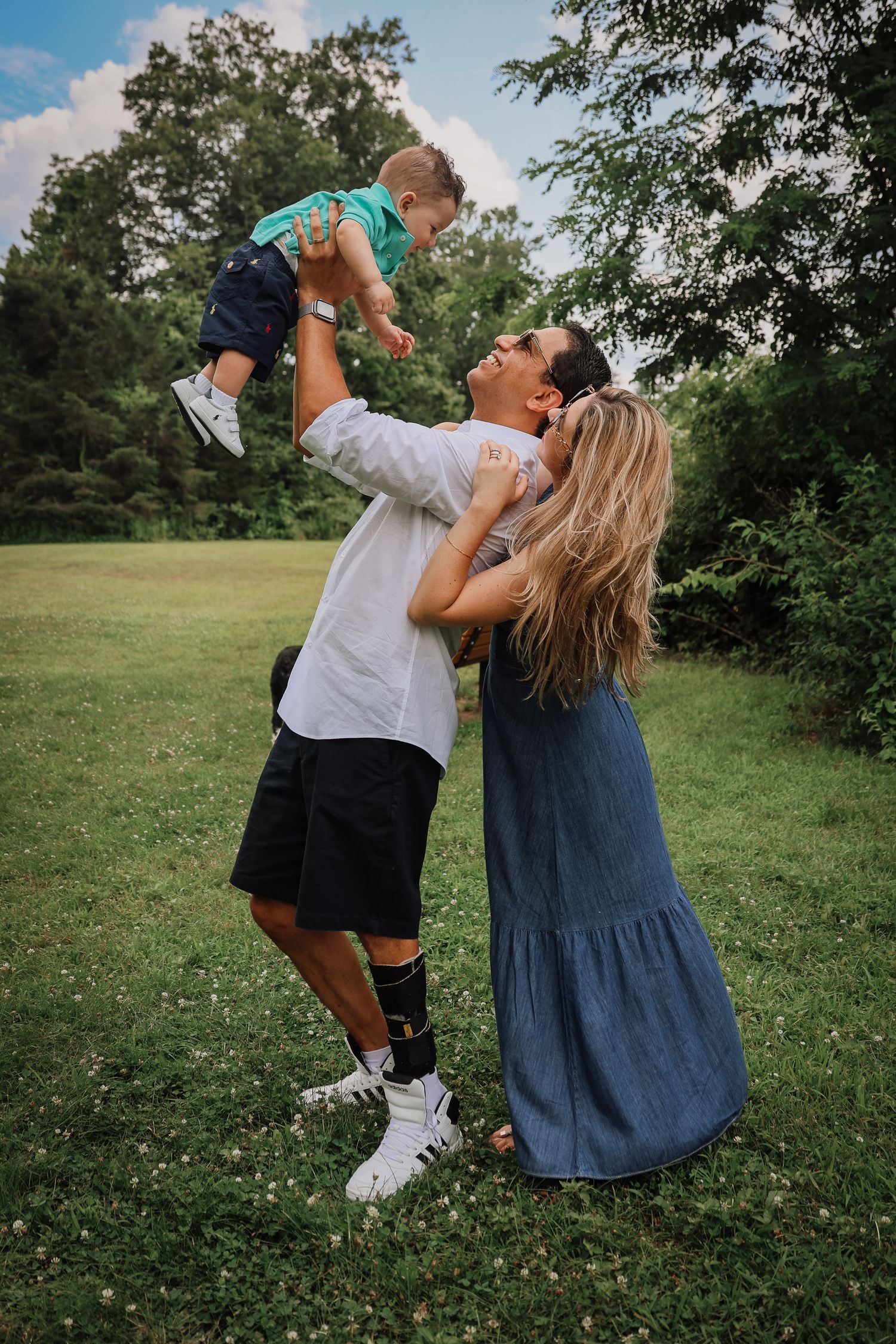A family plays together outdoors in a grassy field on a sunny summer day, lifting a small child up in the air.