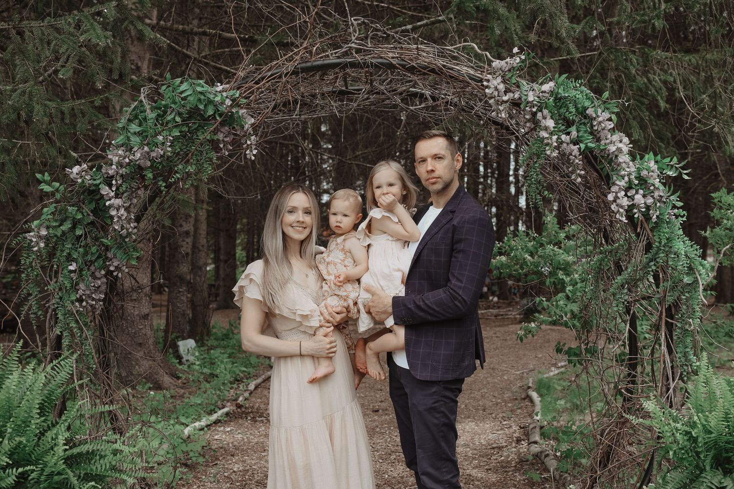 A family poses under a rustic floral arch in a wooded setting wearing elegant neutral-toned attire.