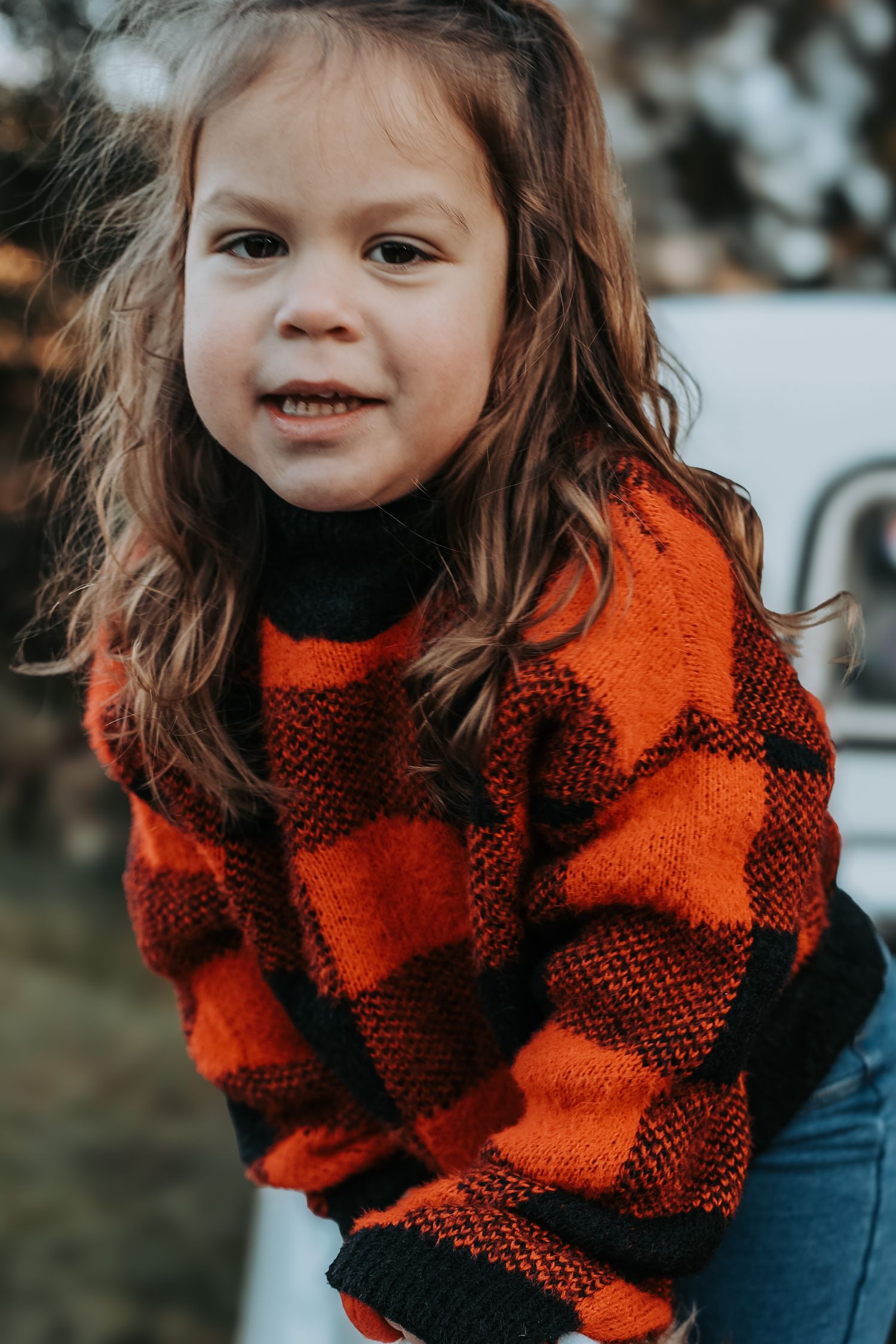A child wearing a cozy red and black buffalo plaid sweater smiles during an outdoor winter photoshoot.