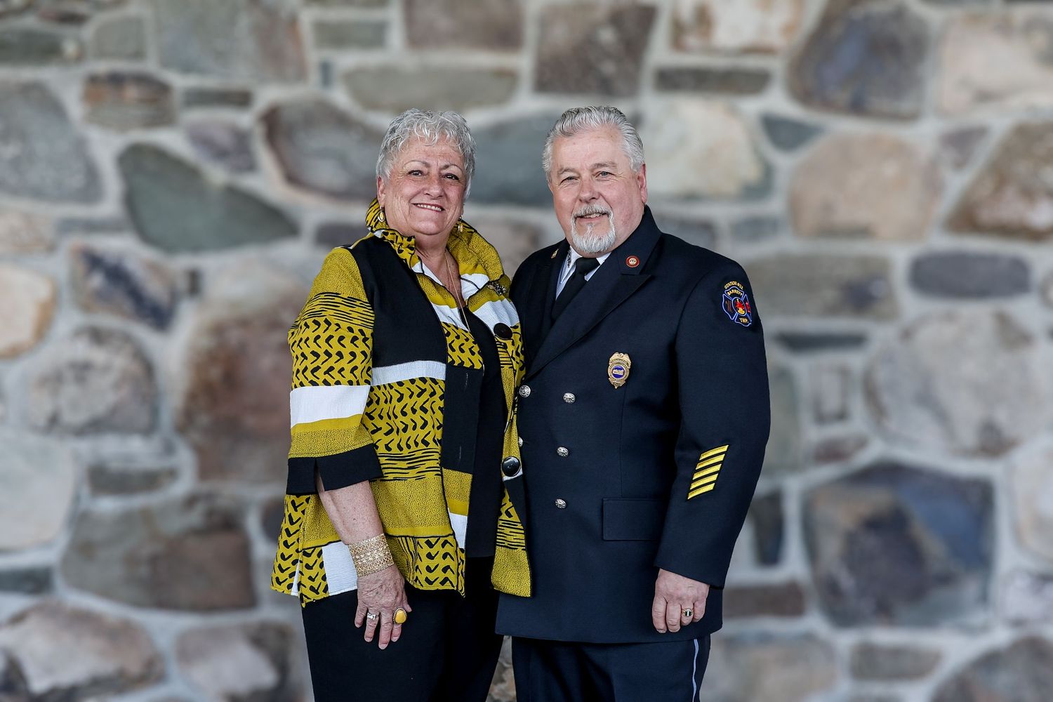 A couple poses together against a stone wall backdrop wearing yellow and black attire.