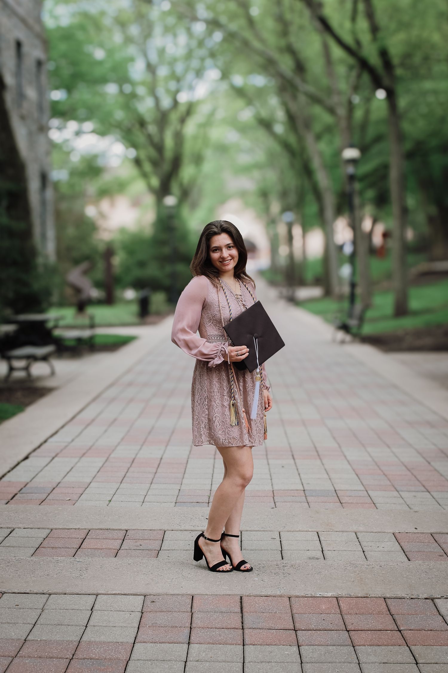 Someone in a pink dress and heels poses with a graduation portfolio on a tree-lined brick pathway at a university campus.