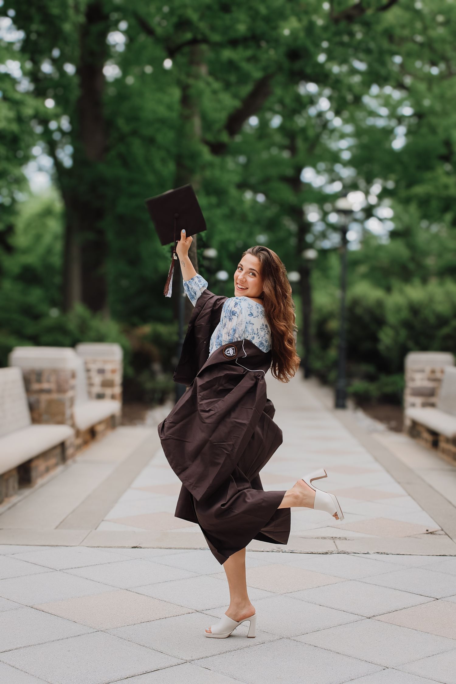 A dancer in a flowing brown dress performs graceful movements on a stone pathway surrounded by green trees.