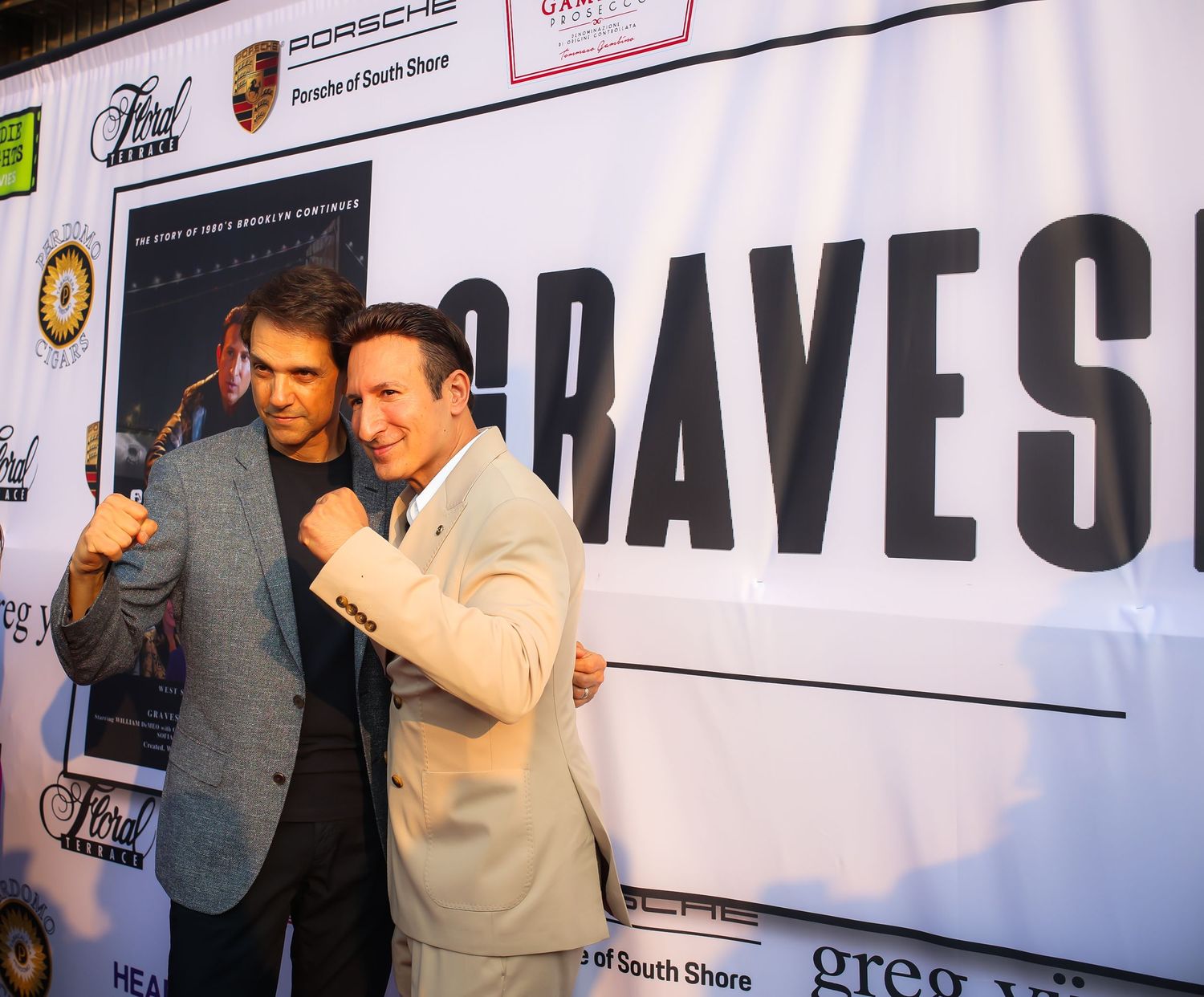 Two men pose together and smile in front of a large banner at a promotional event.
