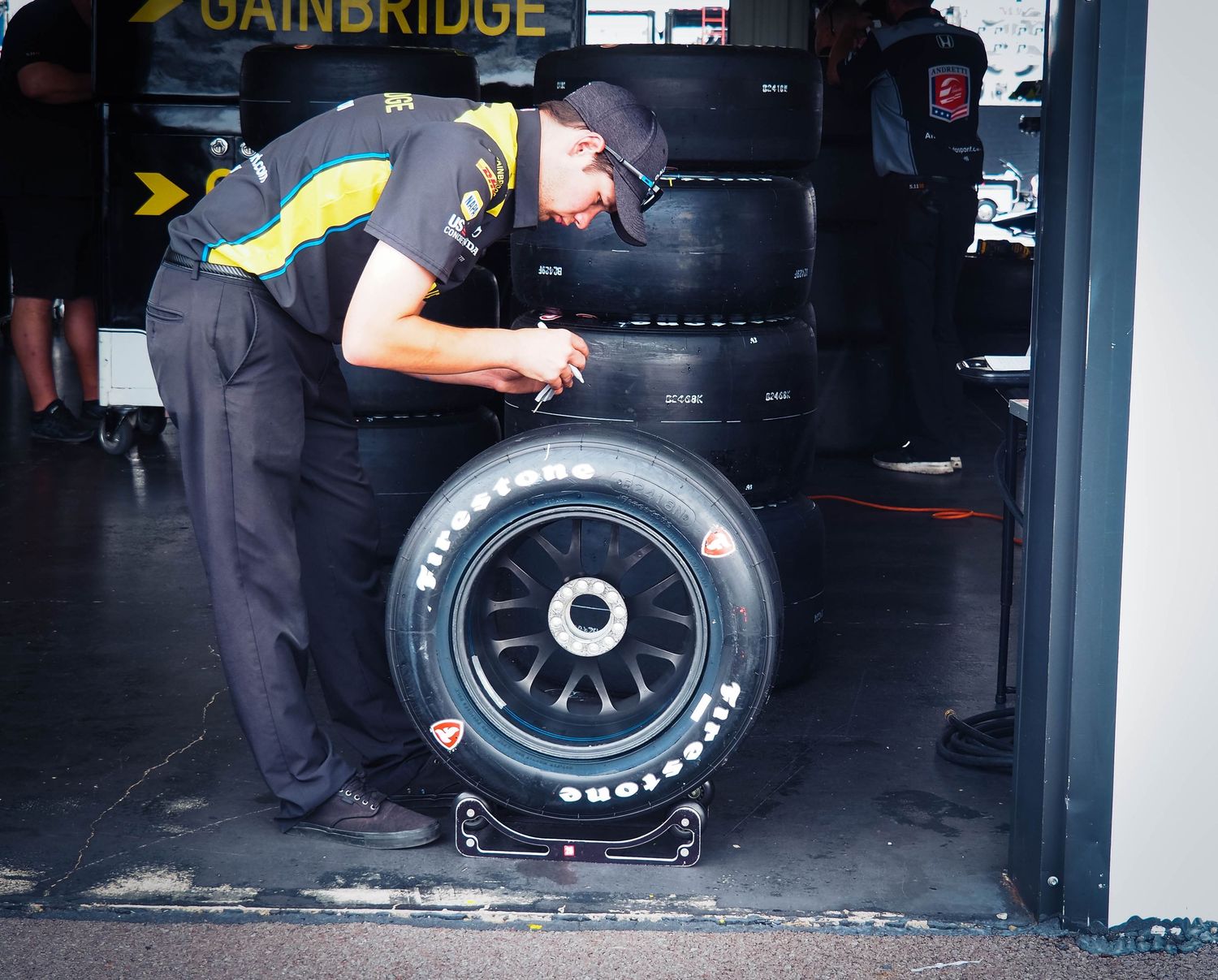 Multiple racing tires are lined up against a dark garage wall with yellow signage visible above.