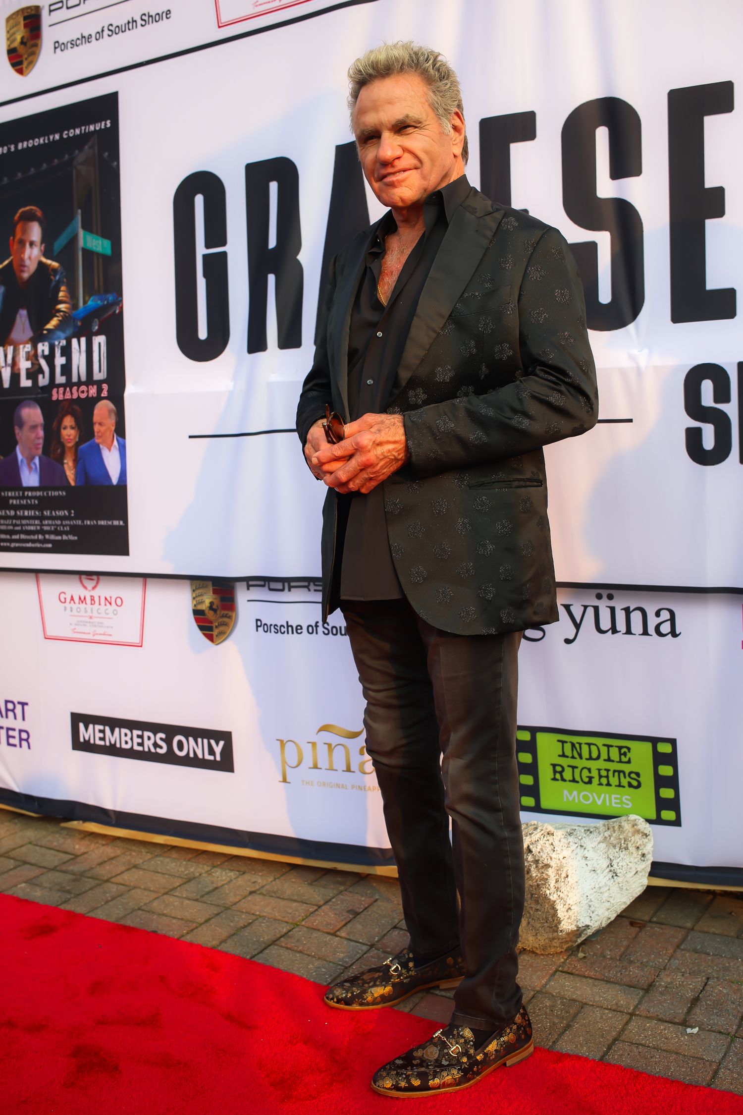 Person in black suit poses on red carpet with promotional banners and posters in background at event.